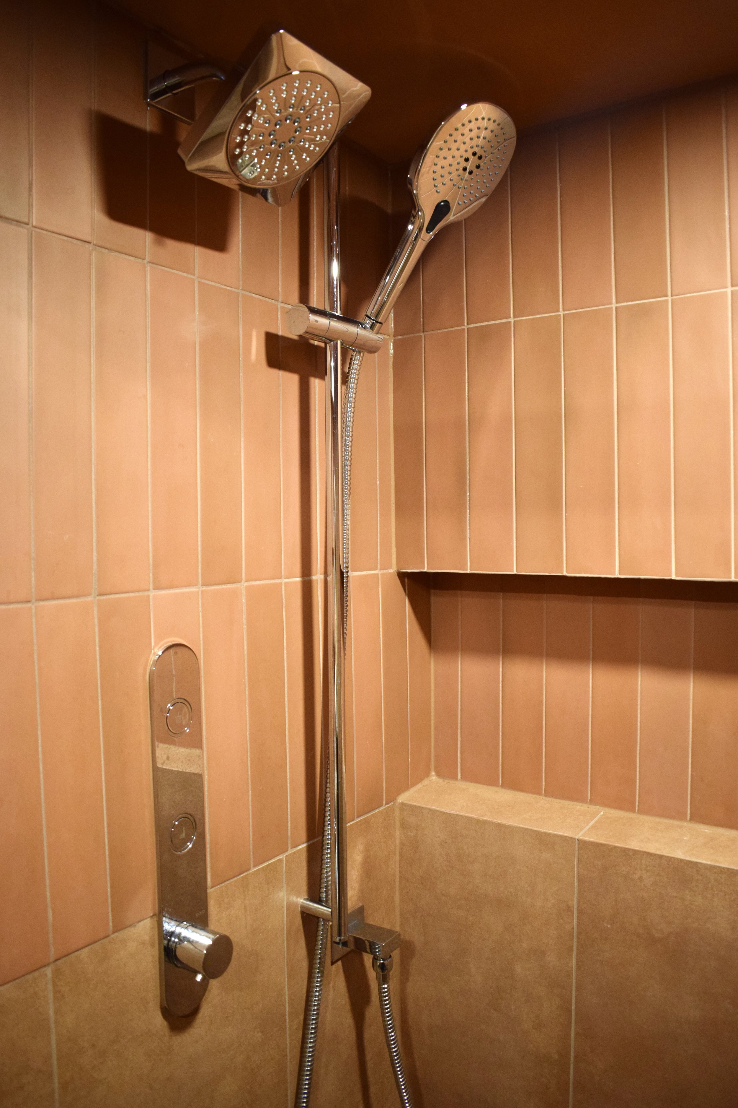 A shower with orange-brown tiled walls, a rainfall showerhead, a handheld showerhead, and a built-in corner seat.