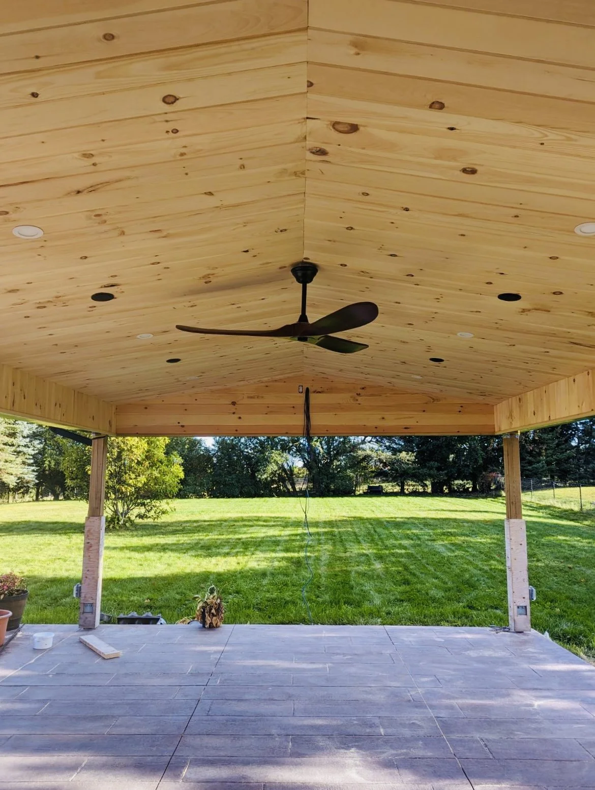 Wood-paneled outdoor ceiling with a ceiling fan in a backyard covered patio, with a view of a grassy yard and trees outside.