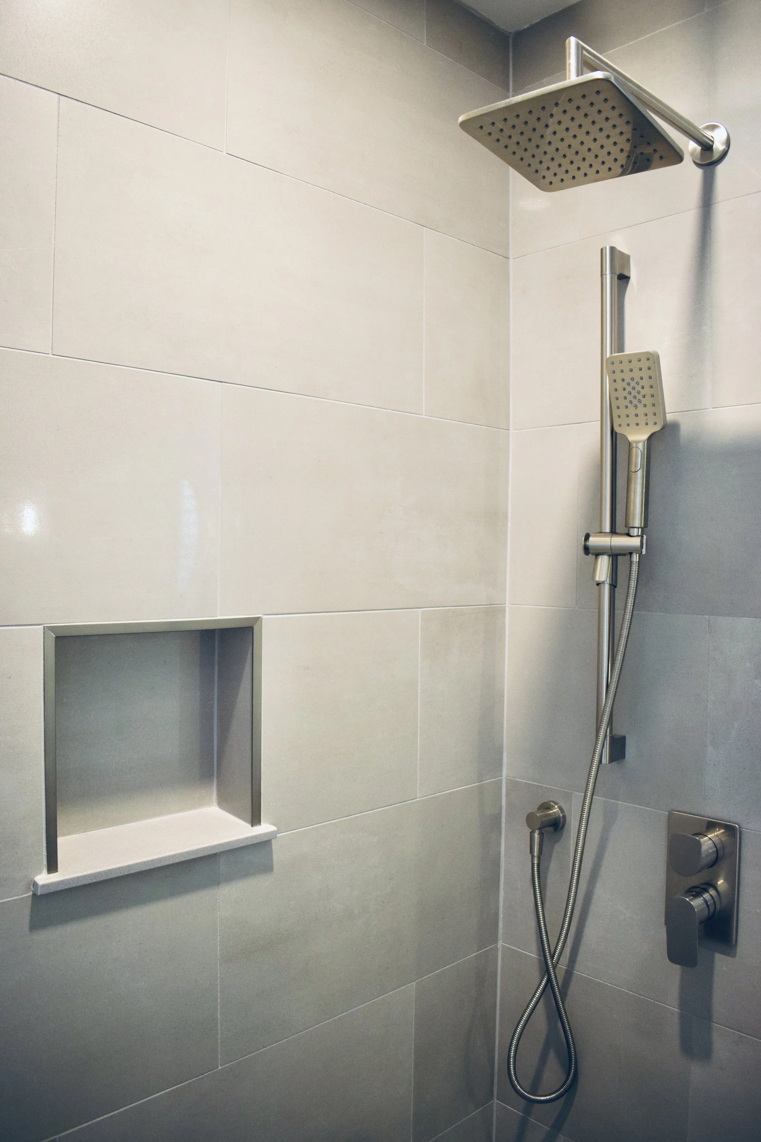 A shower with a large square rain showerhead, a handheld shower nozzle, a modern wall-mounted control panel, tiled walls, and a built-in shower niche.