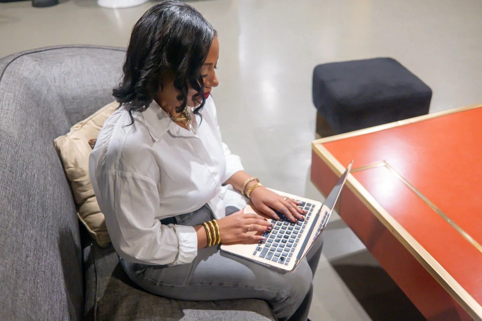 Ariane Lovell - in white blouse, gold jewelry, jeans sitting down typing on laptop on her lap