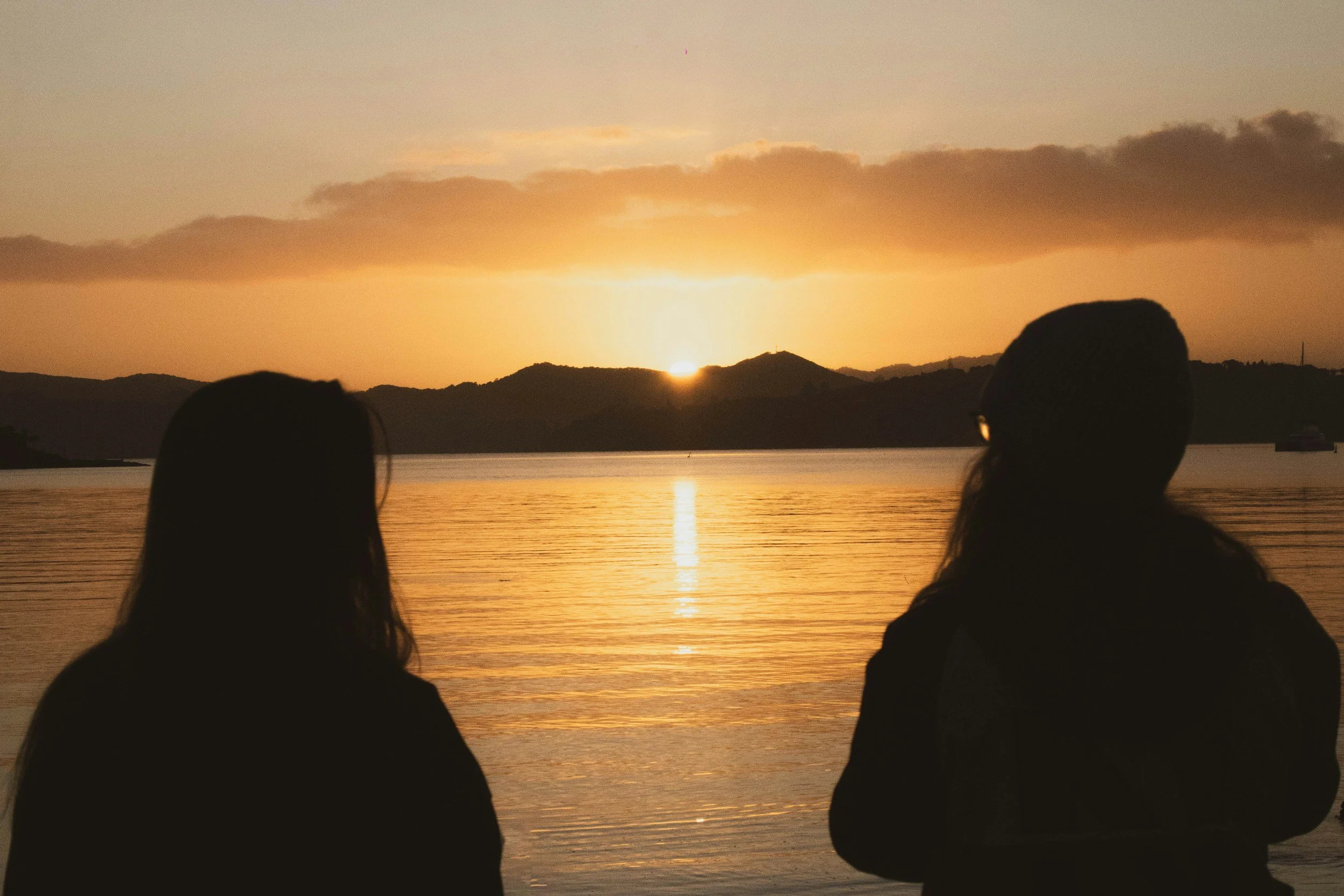 Two silhouetted women watching a sunset over a body of water with hills in the background.