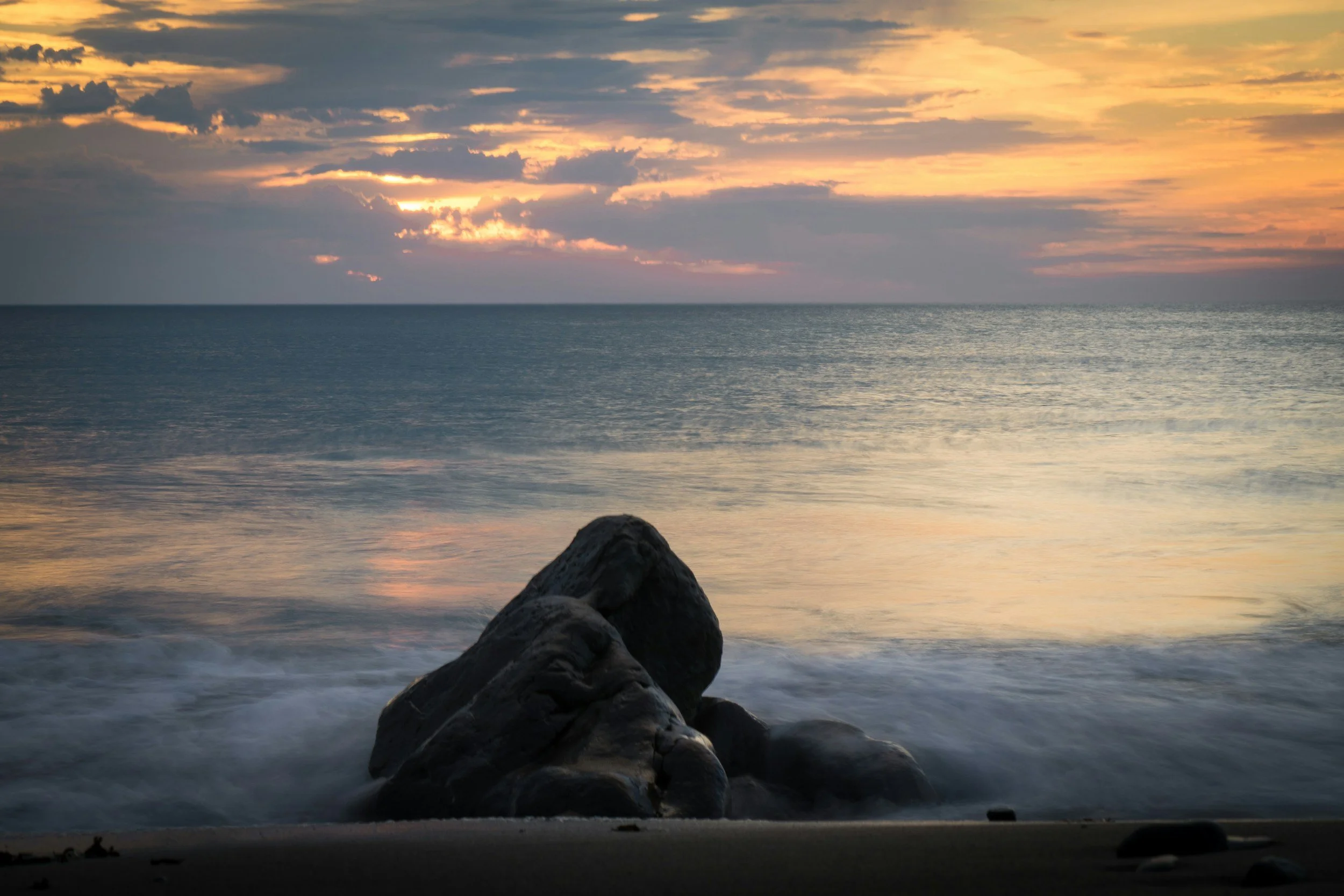 Sunset over the ocean with colorful clouds, waves crashing against rocks on a sandy beach.