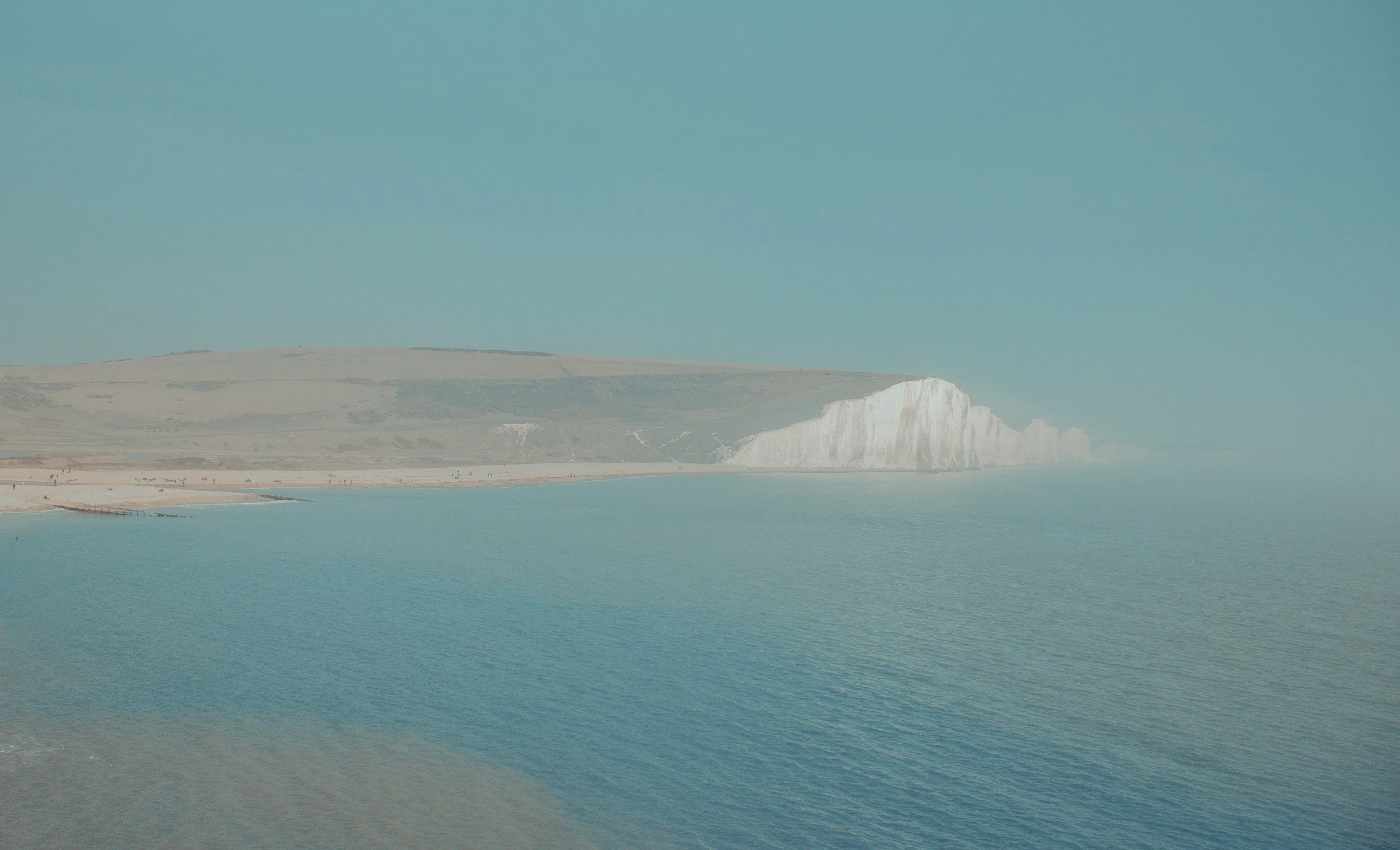 Coastal view of white chalk cliffs, a sandy beach, and calm blue water under a clear sky.