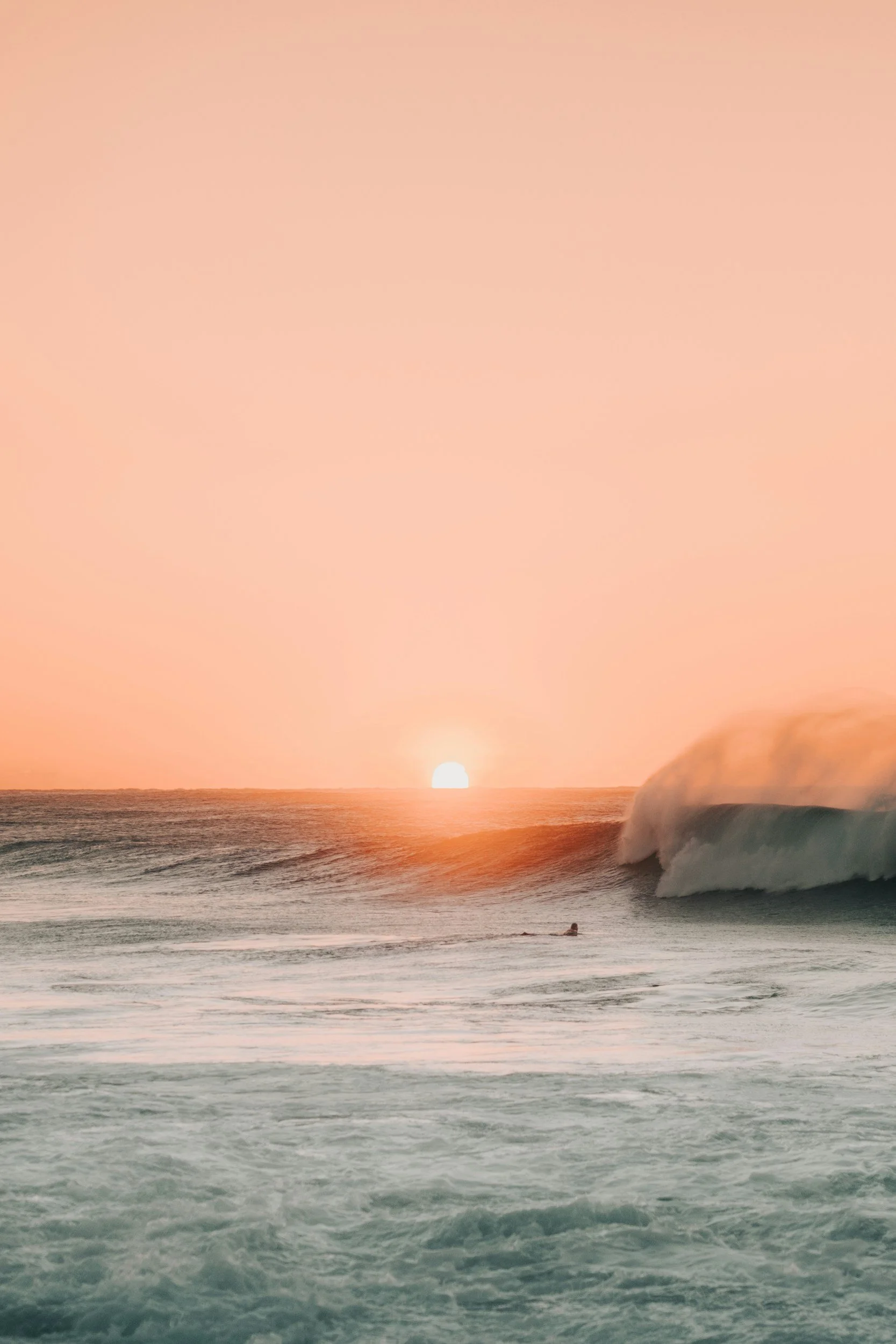 Sunset over the ocean with waves, pink sky, and a surfer in the water.