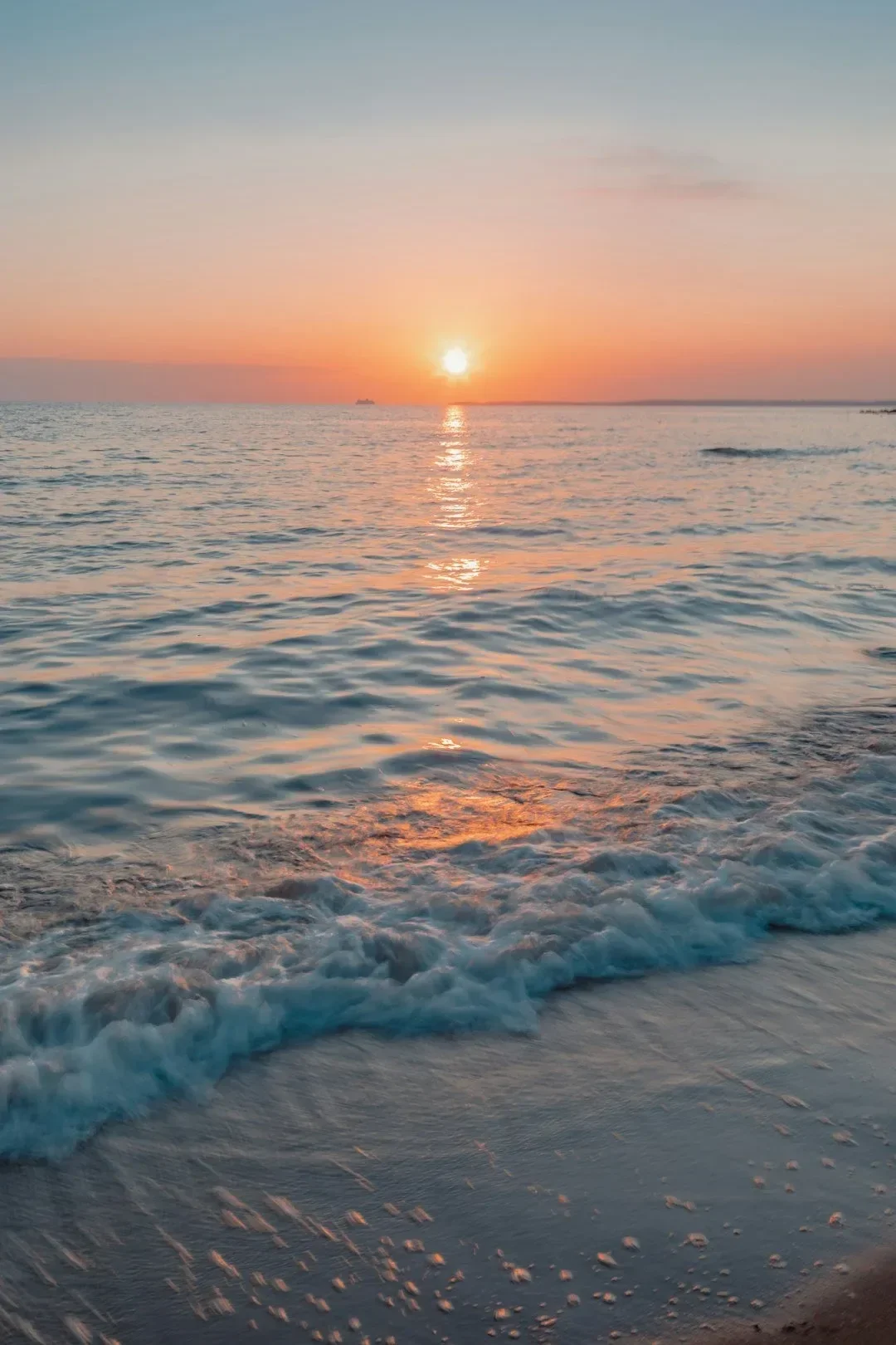 A sunset over the ocean with the sun near the horizon and its reflection on the water, calm waves along the shore, and a ship in the distance.