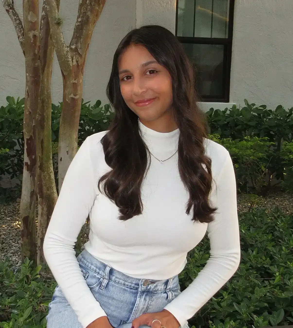 A young woman with long, dark wavy hair wearing a white long-sleeve top and light blue jeans, smiling while standing outdoors near a tree and green bushes.