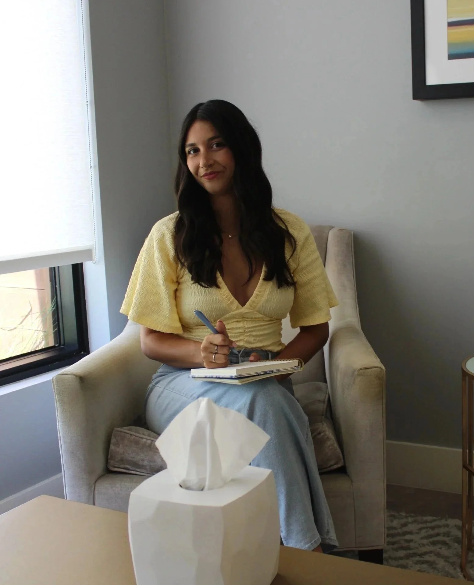 A woman with long dark hair, wearing a yellow top and light blue jeans, sitting on a beige armchair next to a window with white blinds, holding a notebook and pen, with a tissue box on a side table in front of her.