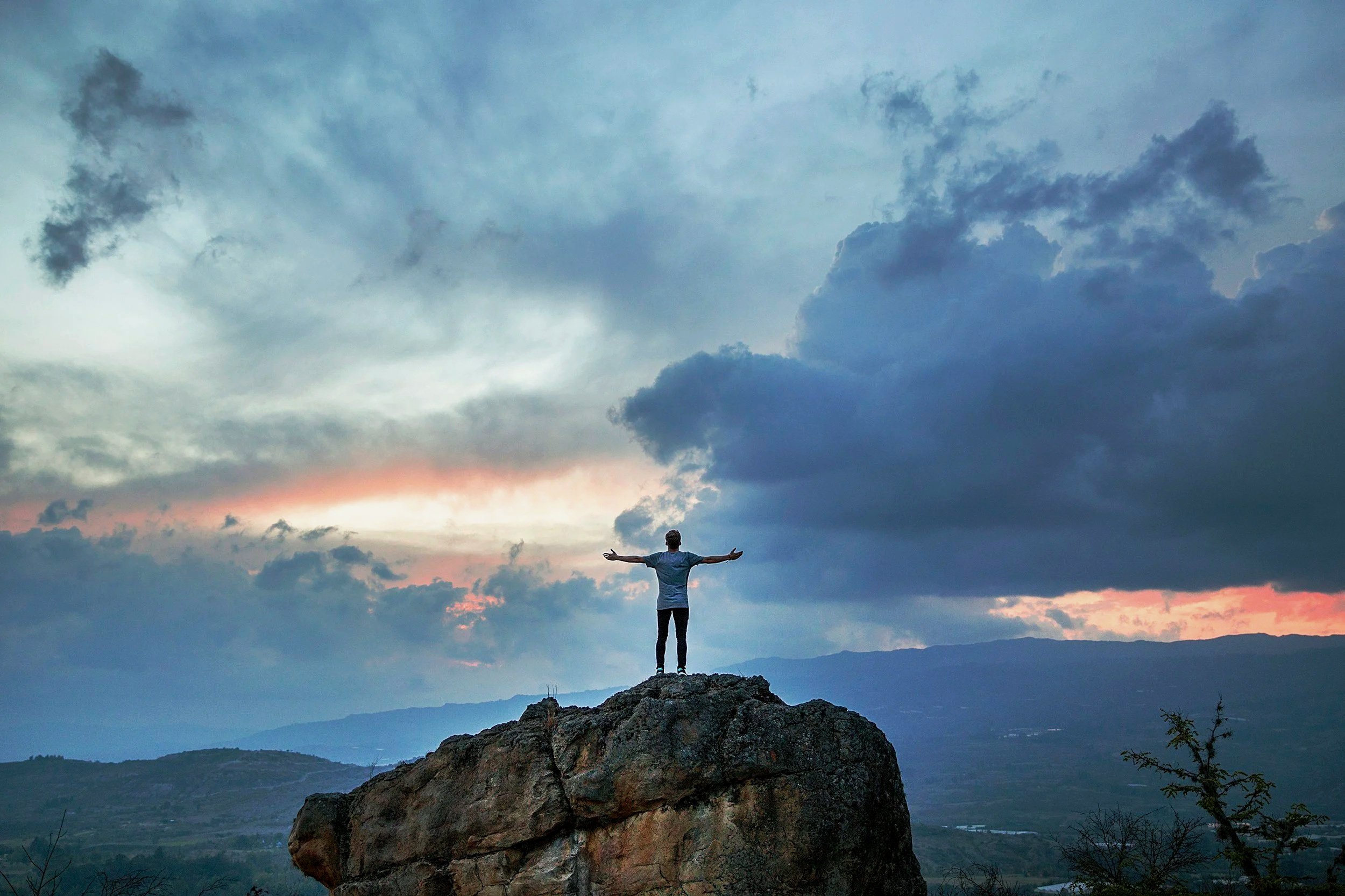 a picture of a man with his arms spread open as he stands on a precipice of a mountain overlooking a mountain range at dusk