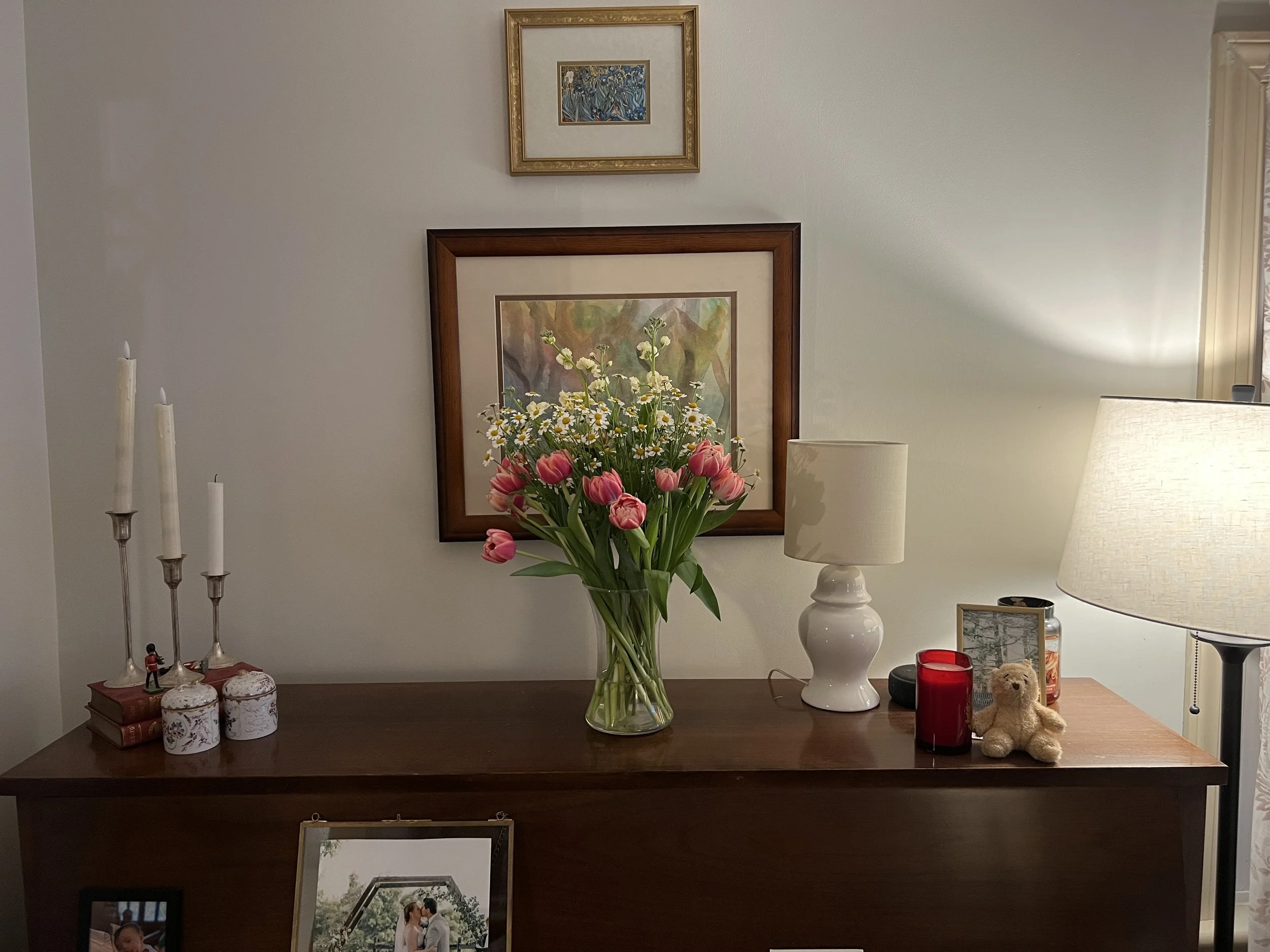 A wooden table with a large glass vase filled with pink tulips and small white flowers in the center. To the left, there are three white candles in silver holders and small decorative boxes. To the right, there is a white table lamp and a smaller lam