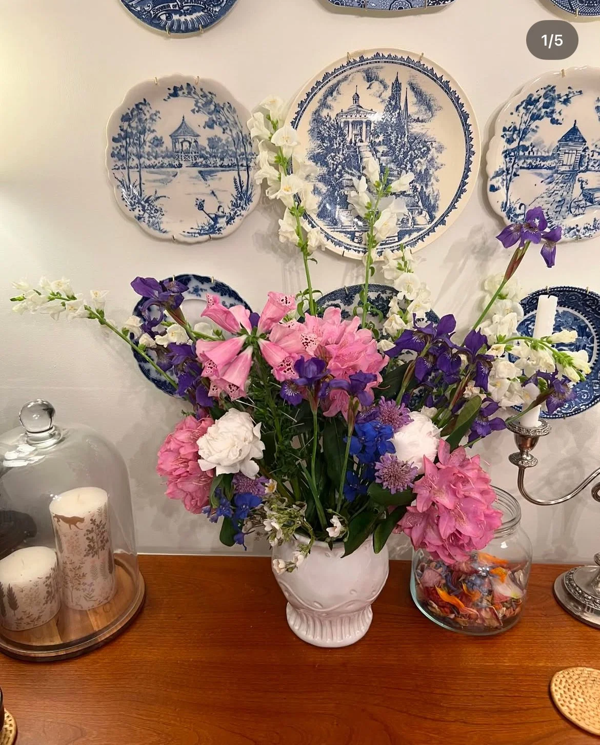 A large bouquet of pink, white, purple, and blue flowers in a white vase on a wooden table, with decorative blue and white plates on the wall behind it, and candles and other decorative items around.