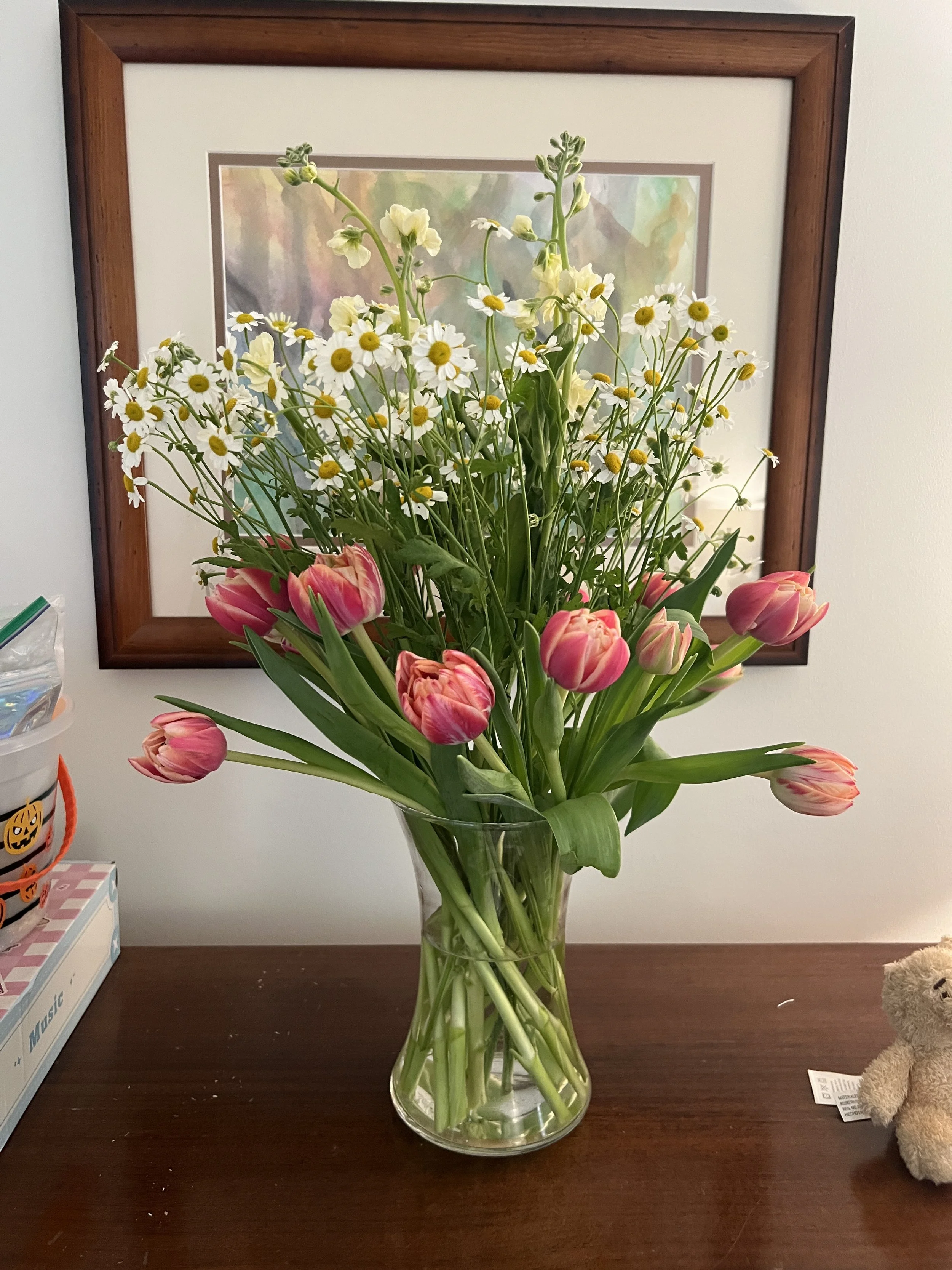 A clear glass vase holding a bouquet of pink tulips and white daisies on a wooden surface, with a framed picture on the wall behind.
