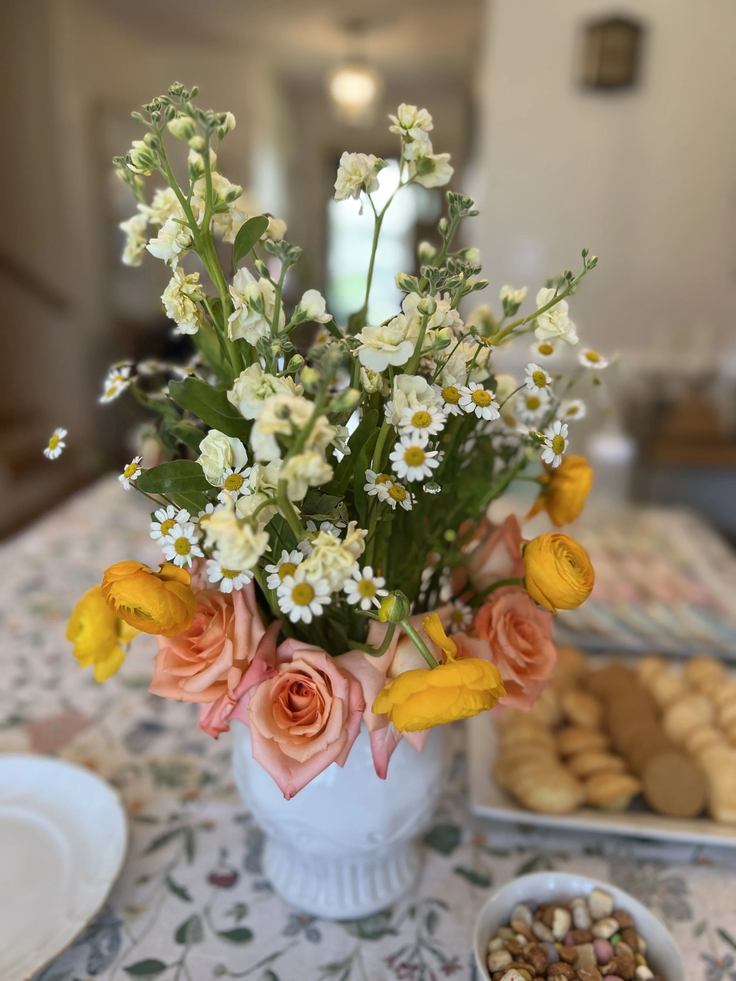 A floral arrangement with pink roses, yellow ranunculus, white daisies, and white stock flowers in a white vase, placed on a table with a floral tablecloth.