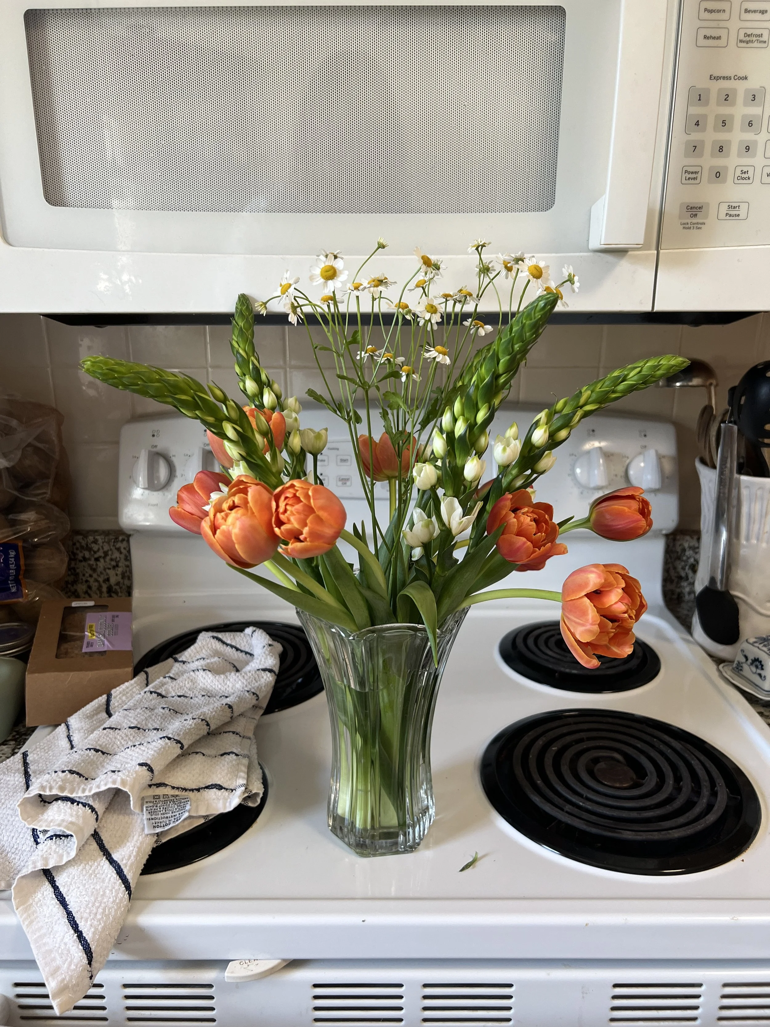 A glass vase with orange tulips and white daisies placed on a white stove in a kitchen.