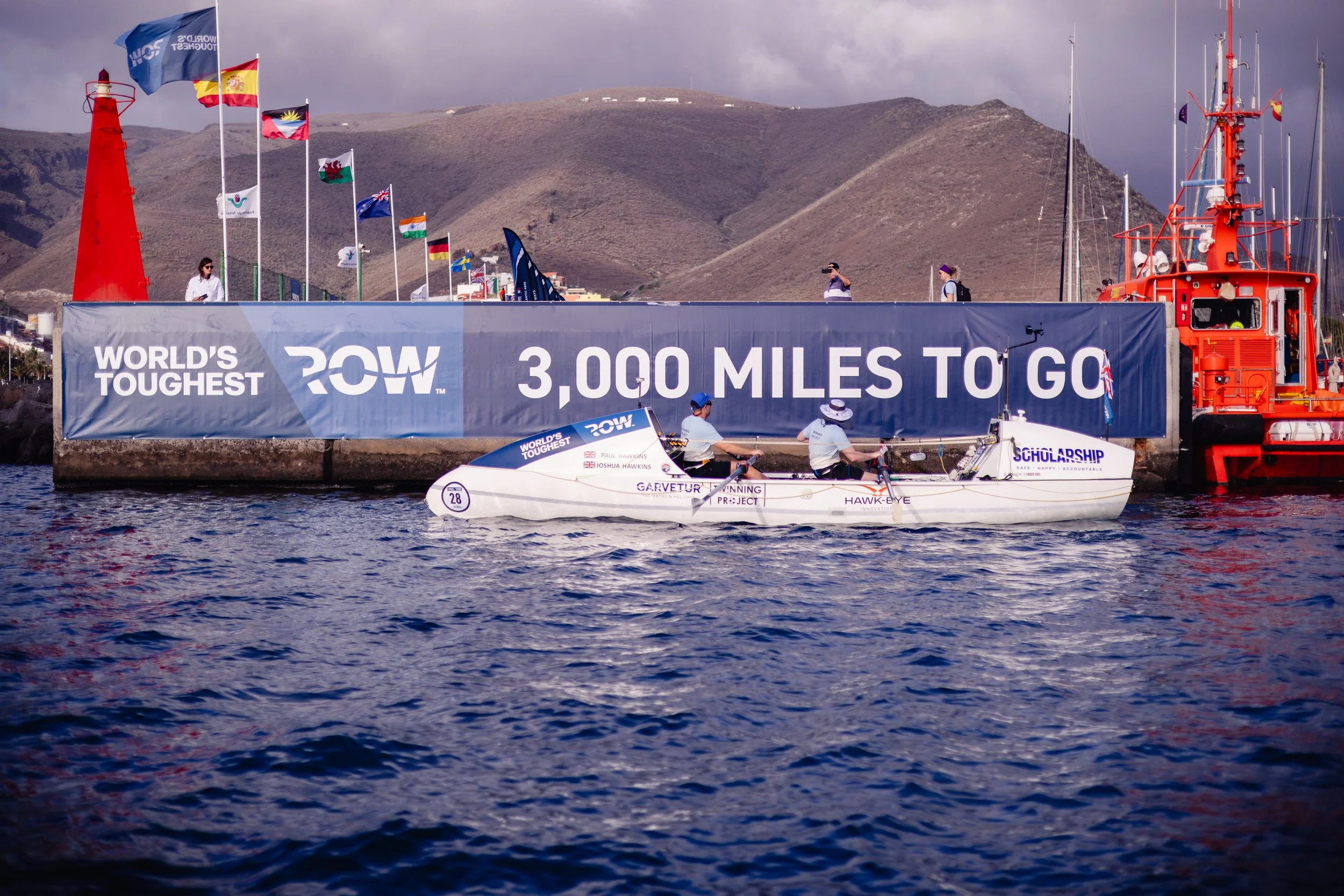 A sailing boat passing by a harbor with a large banner advertising 3,000 miles to go in a race, with flags flying and hills in the background.