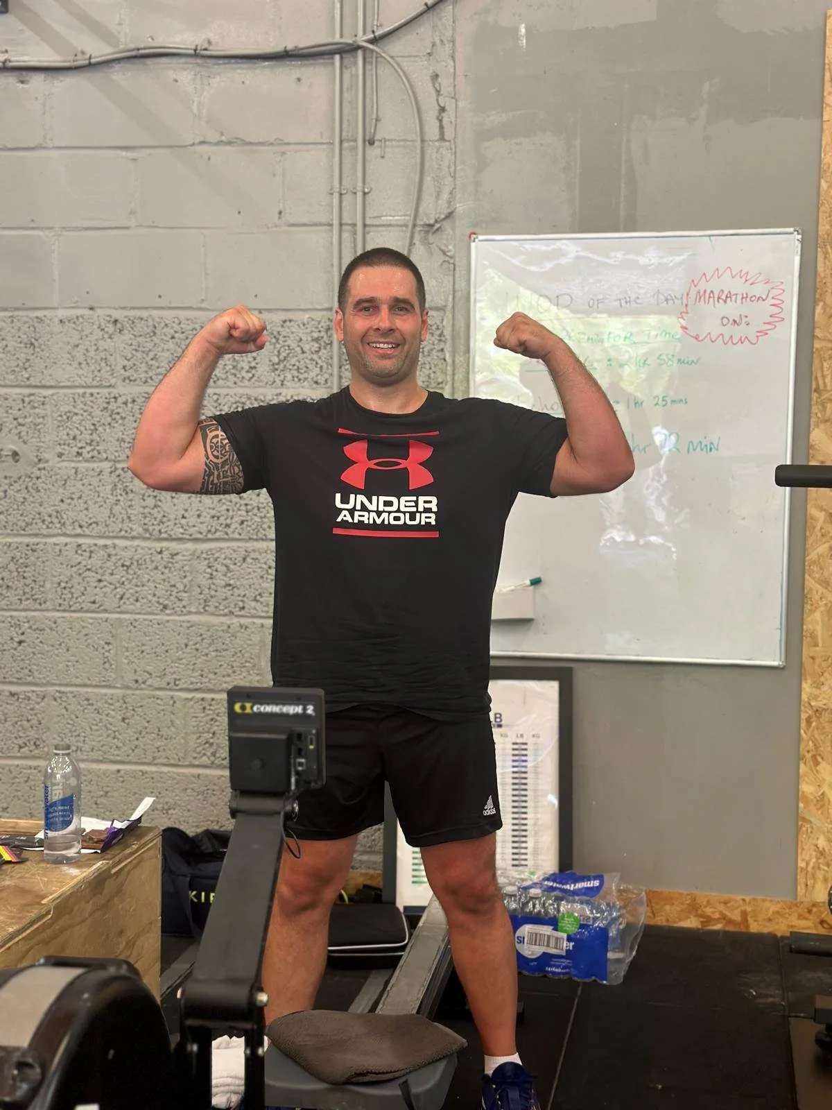 Man in a black Under Armour t-shirt and shorts flexing his biceps in a gym or training room, smiling, with a whiteboard and water bottles in the background.