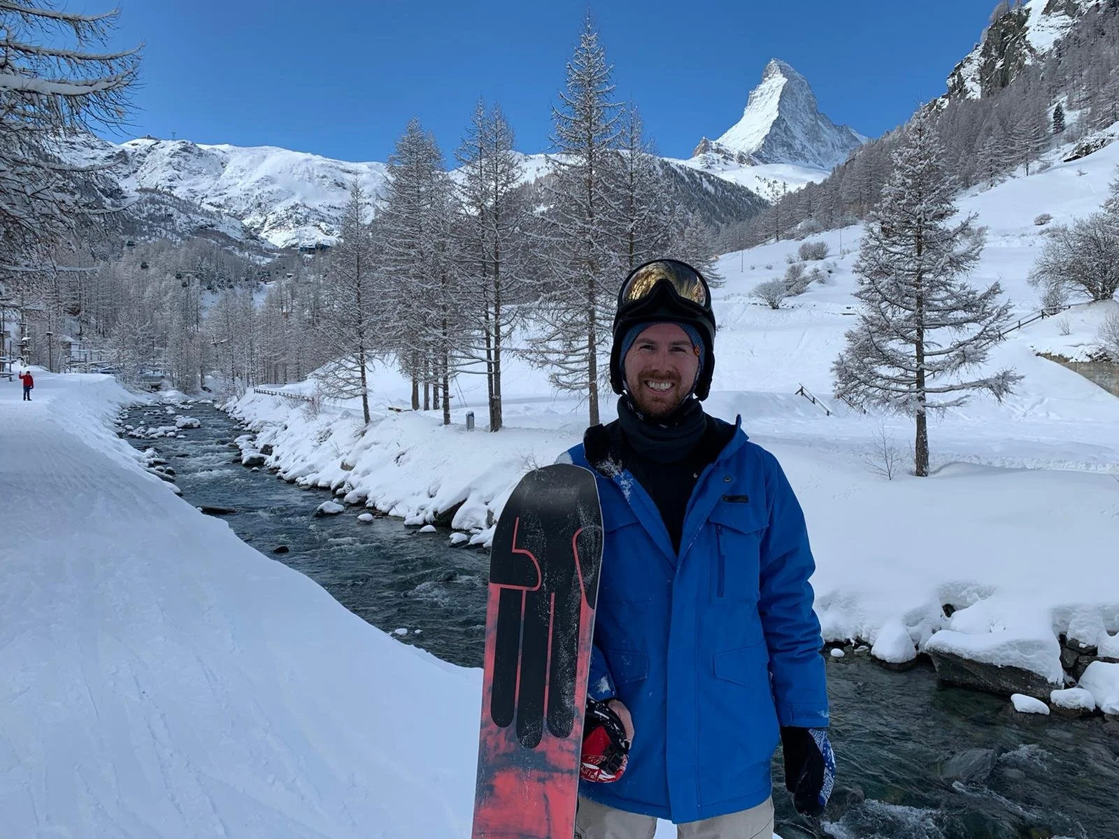 A man dressed in winter gear standing by a snow-covered river in a mountainous landscape with snow-covered trees and a tall peak in the background.