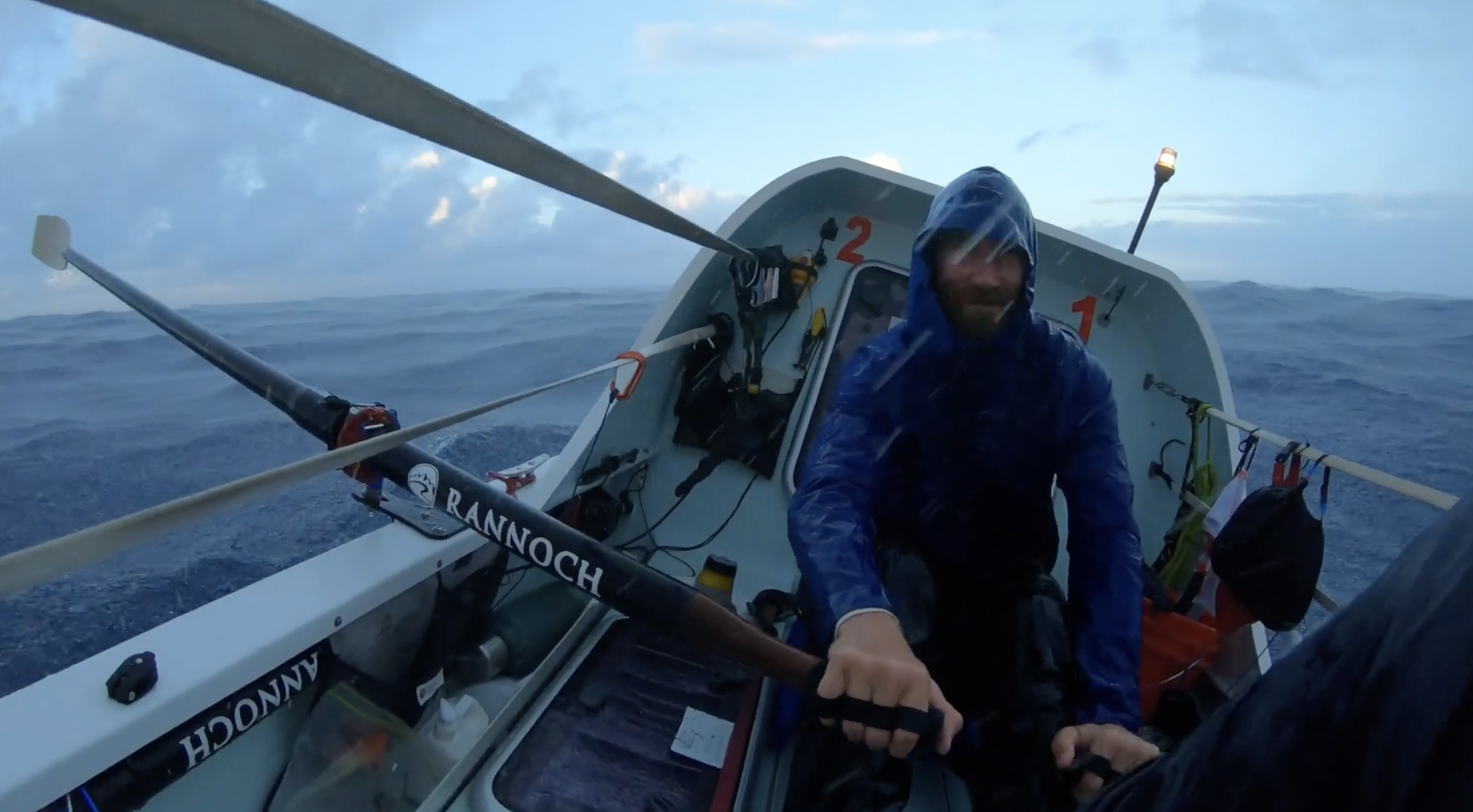A man wearing a blue rain jacket and hood smiling while sitting in a small boat in rough ocean waters during a rainy day, holding a paddle labeled 'RANNOCH' with life gear and equipment around him.