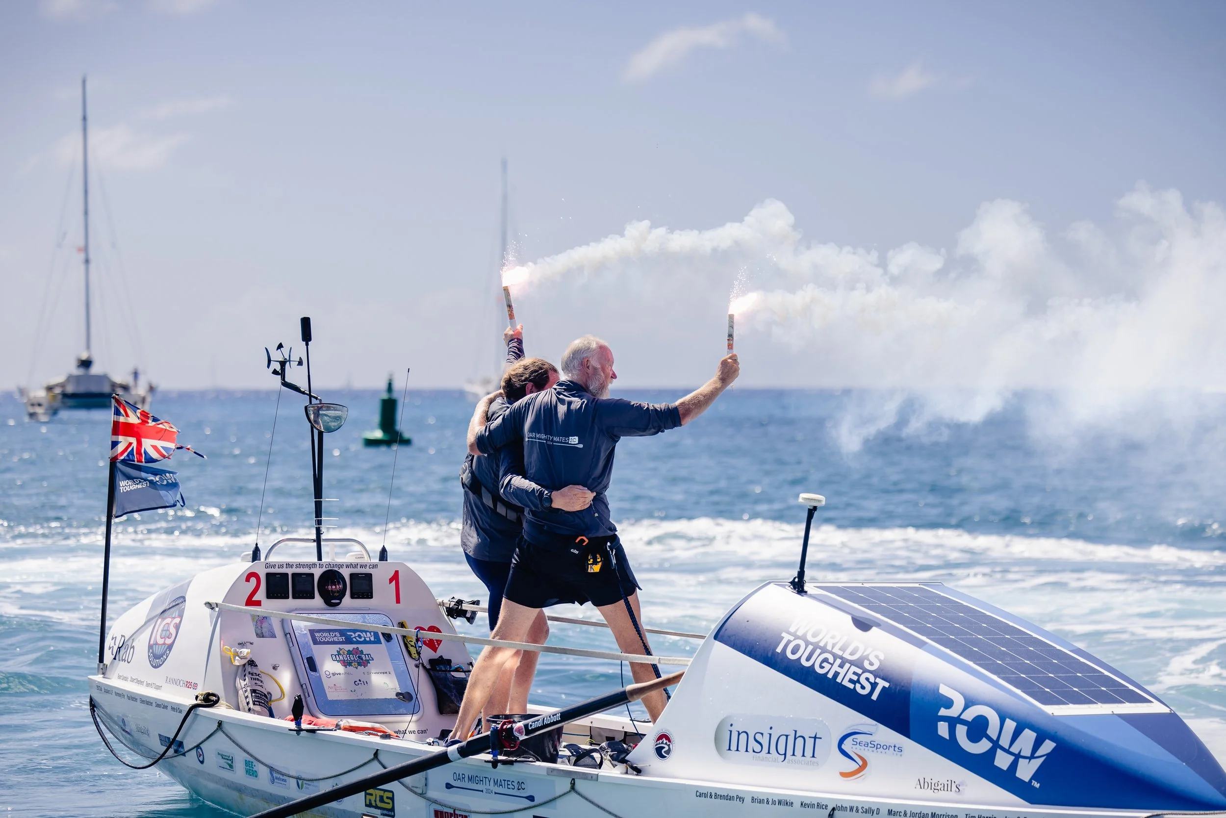 Three people celebrating on a racing boat, holding sparklers with a sea and sailboats in the background.