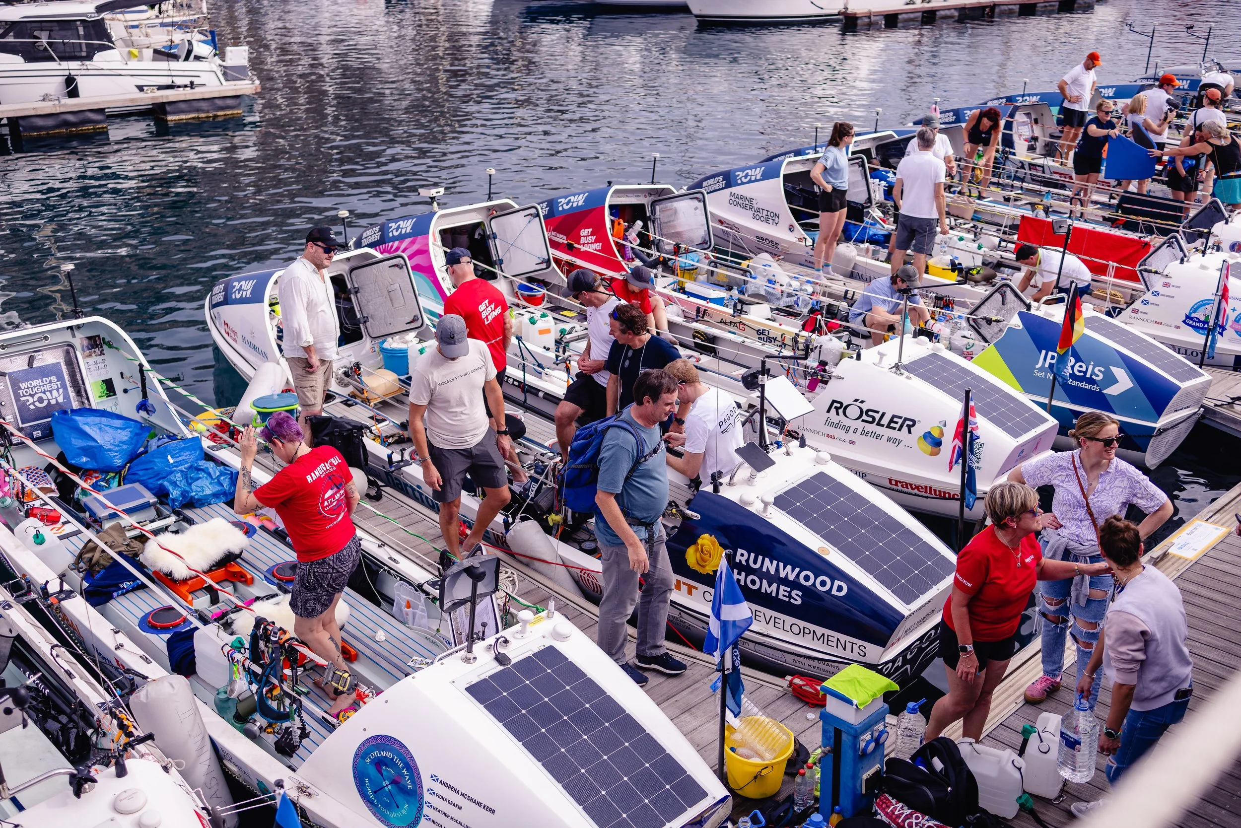 Multiple ocean rowing boats docked at a marina with people preparing and working on the boats, some wearing red shirts, others in casual clothing, with various supplies and equipment on the decks, and water in the background.