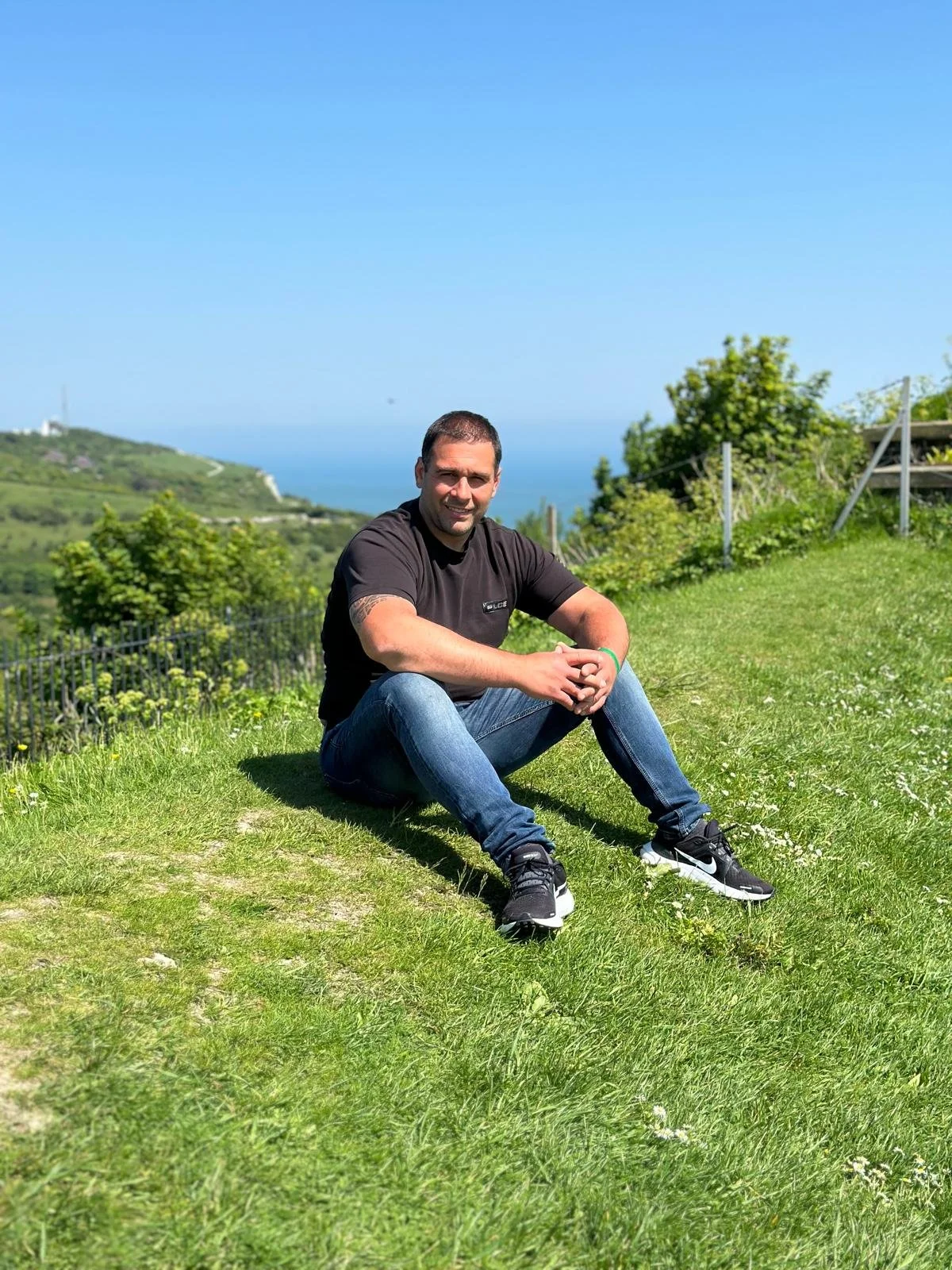 A man sitting on a grassy hill with a scenic view of green hills and the ocean in the background on a sunny day.