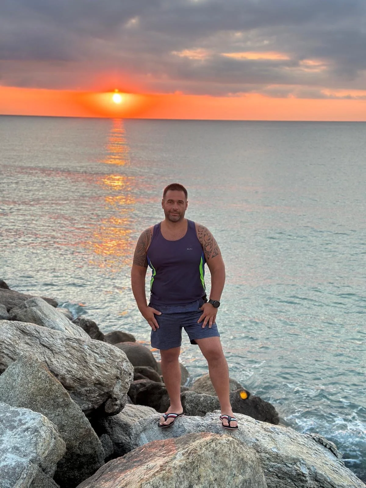 A man standing on rocks by the ocean with a sunset in the background.