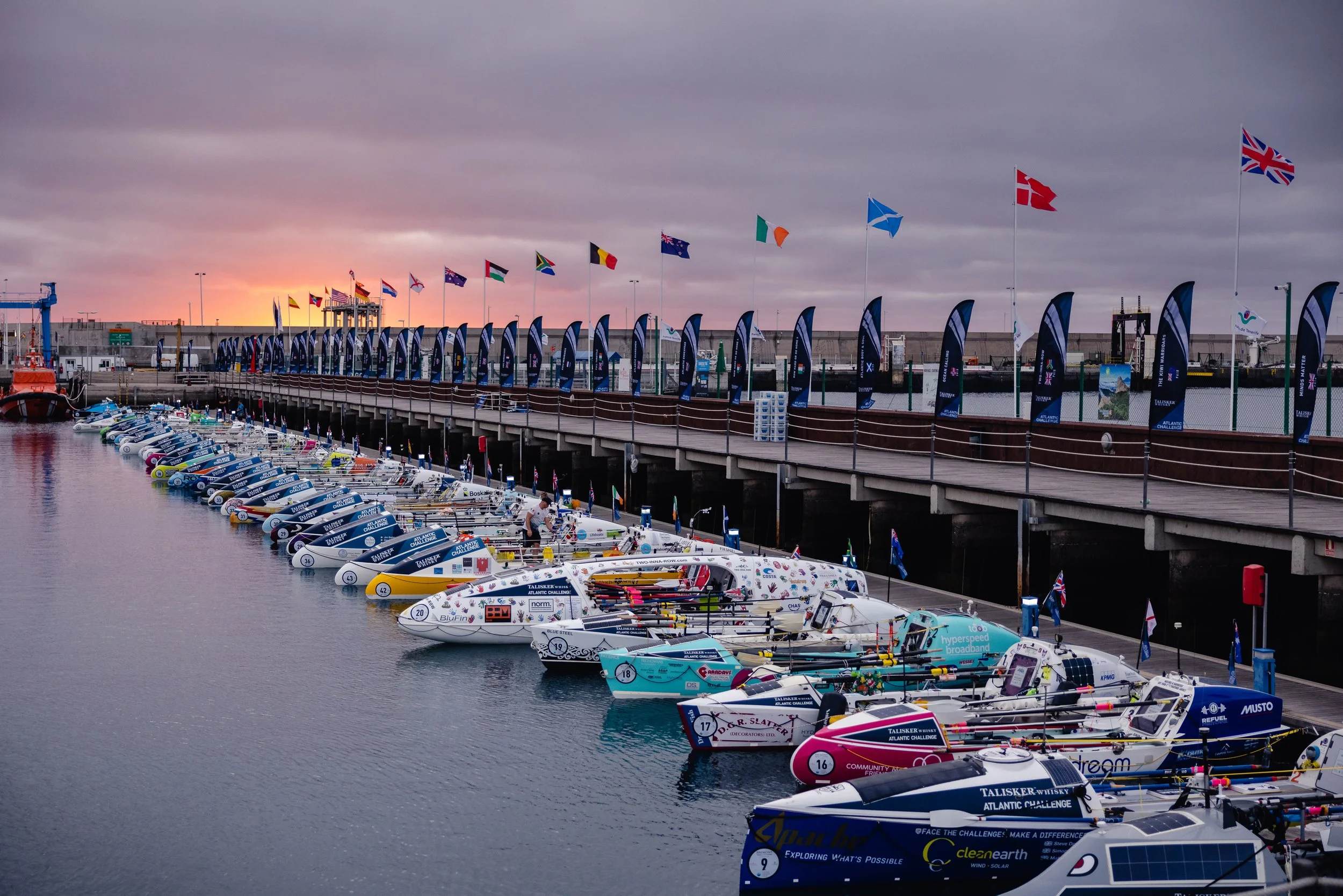 Row of competitive ocean rowing boats docked at a marina during sunset, with flags from various countries flying overhead.
