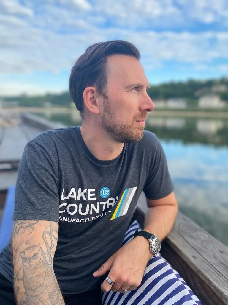 A man with dark hair and a beard sitting outdoors near a lake, wearing a dark t-shirt with a logo, striped pants, and a watch.