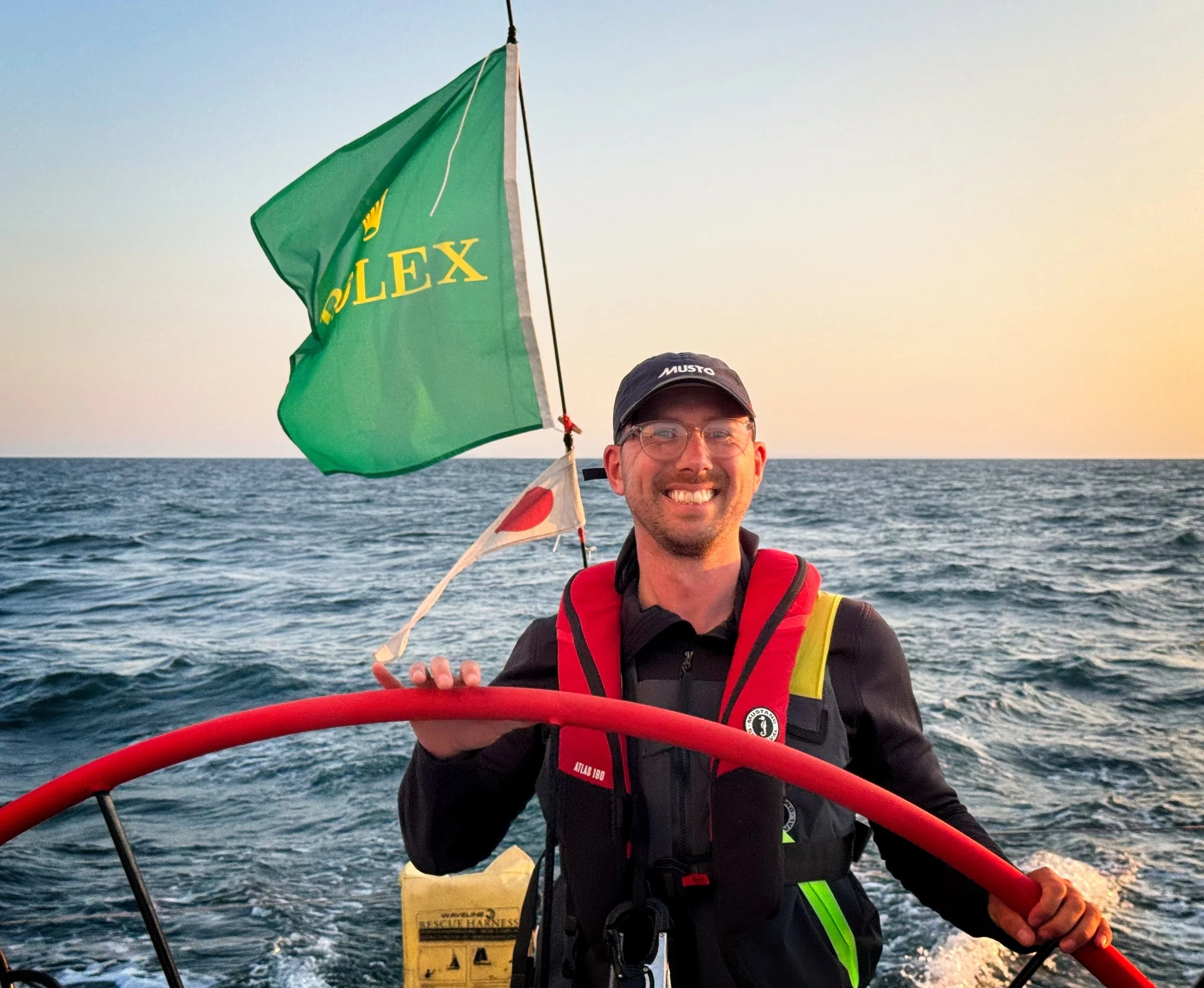 Man sailing on the ocean, smiling, holding a red bar, with a green Rolex flag behind him, during sunset.