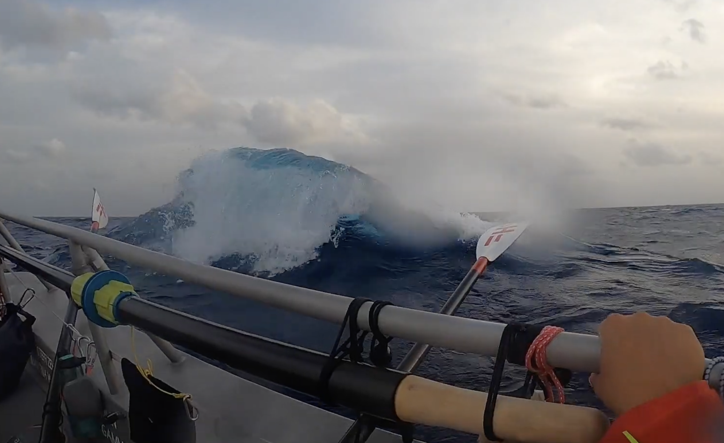 View from a boat showing a large ocean wave about to break, with part of the boat's side and a paddle visible and a person's hand holding another paddle in the foreground.