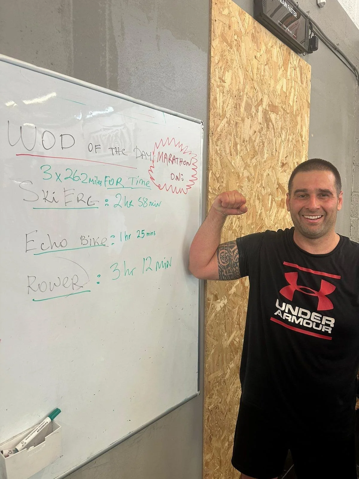 A man with short hair smiling and flexing his right arm, standing next to a whiteboard with handwritten notes about workouts.