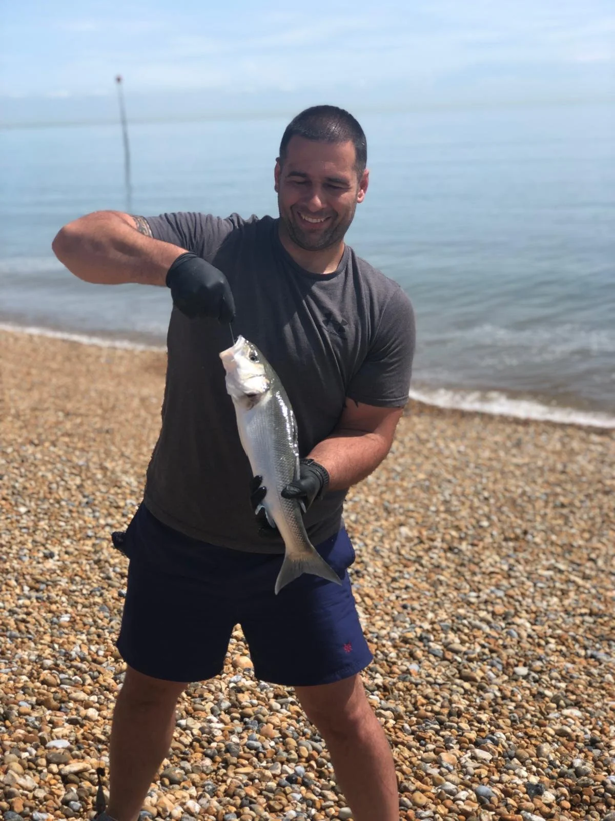 Man holding a fish on a pebble beach near the ocean, smiling.