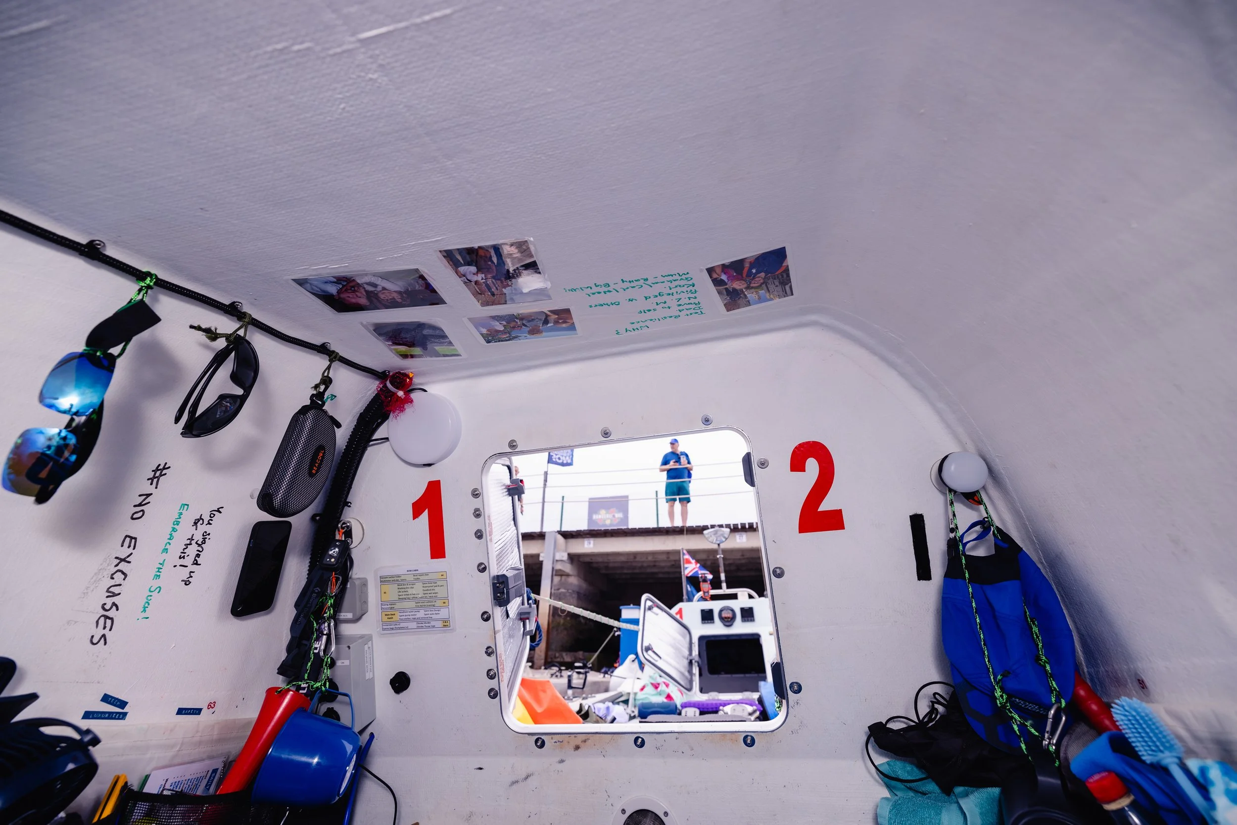 Interior of a boat cabin with equipment hanging on the wall, photographs on the ceiling, and a window showing a person on the dock outside.