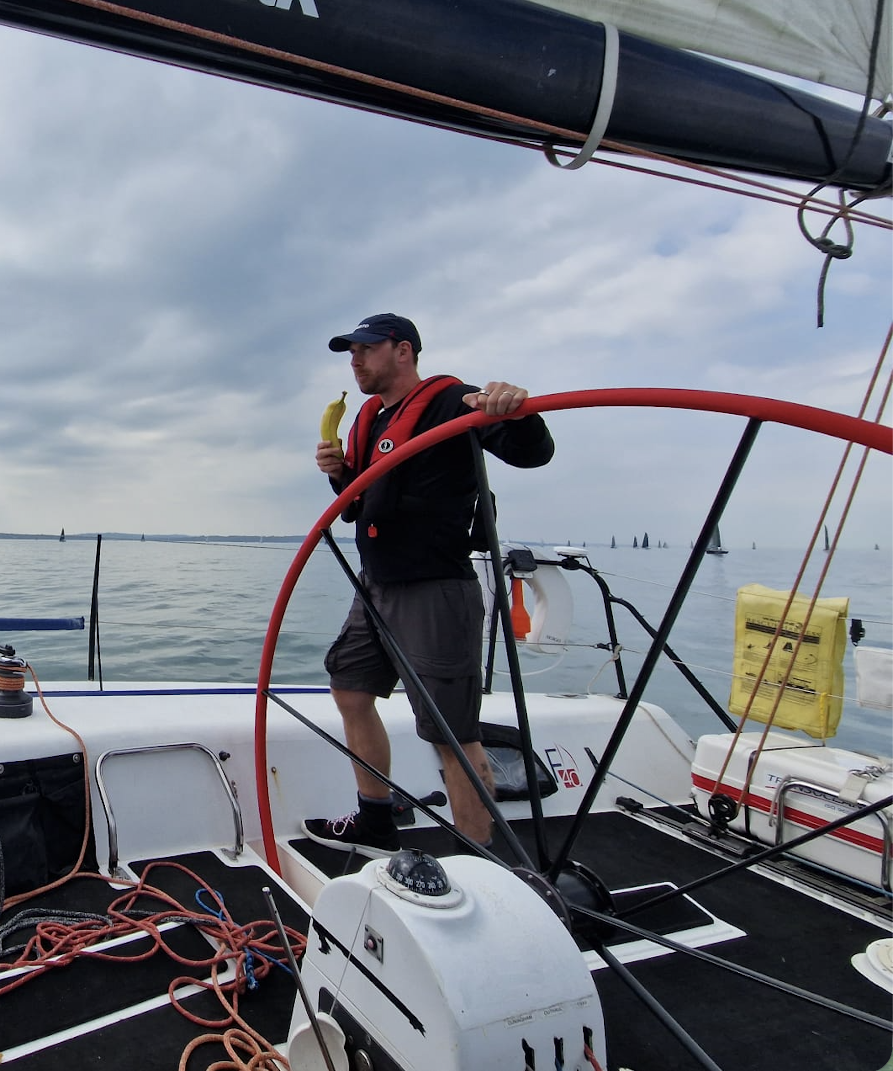 A man standing on a sailboat holding a banana, wearing a black jacket, shorts, and a cap, with a cloudy sky and other sailboats in the background.