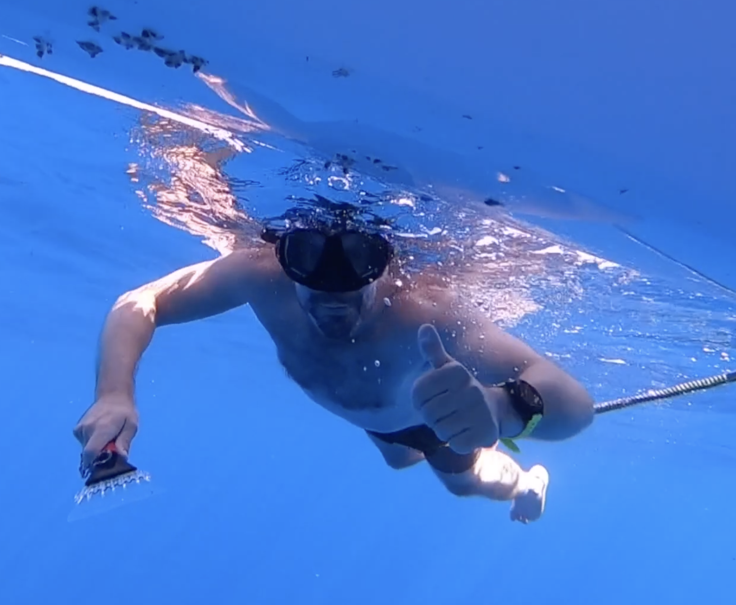 A man swimming underwater in a blue pool, wearing goggles and swimming trunks, giving a thumbs-up sign.