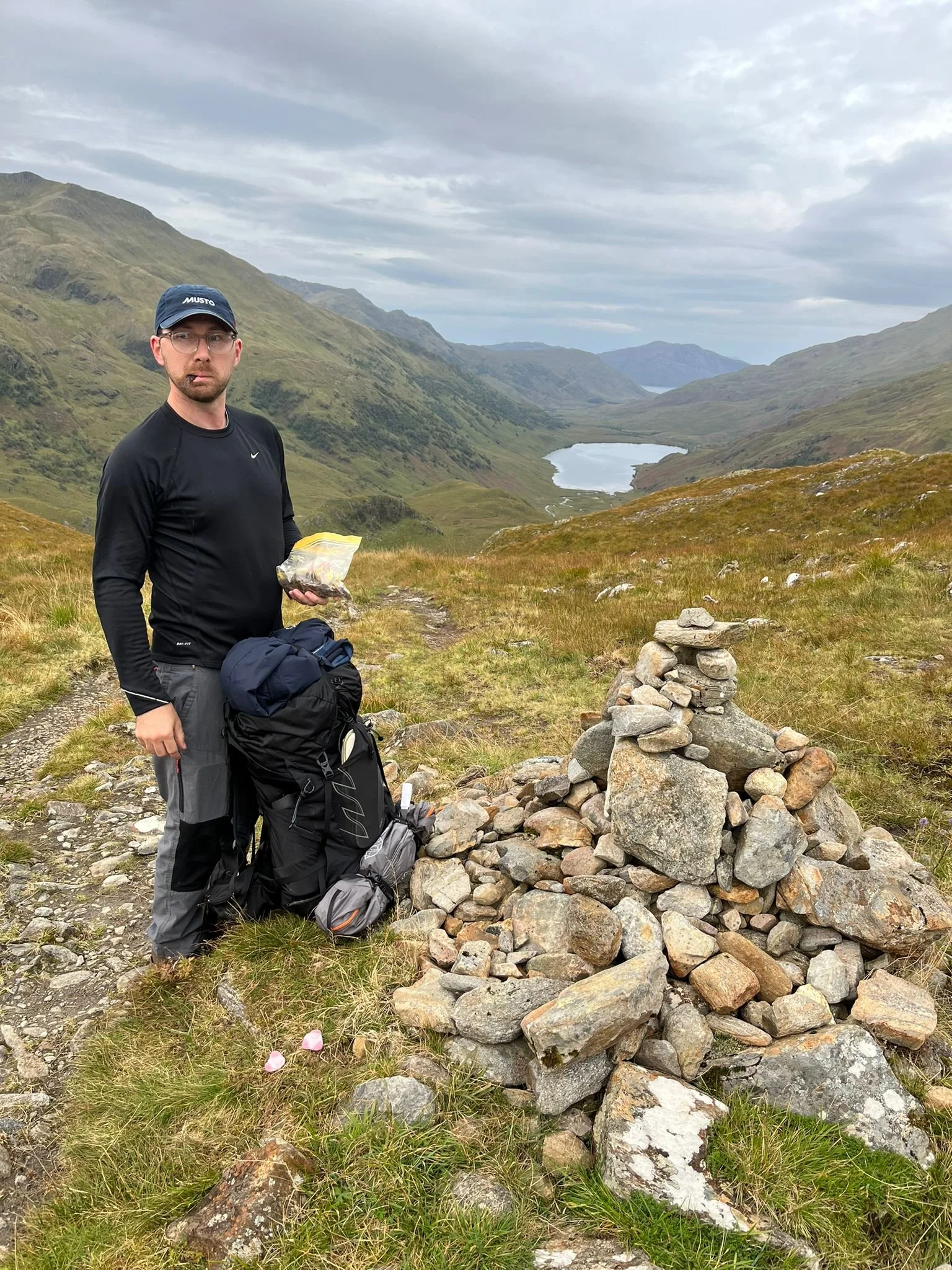 A man standing on a mountain trail with a backpack and holding a snack, next to a cairn of rocks, with green hills, a lake, and cloudy sky in the background.