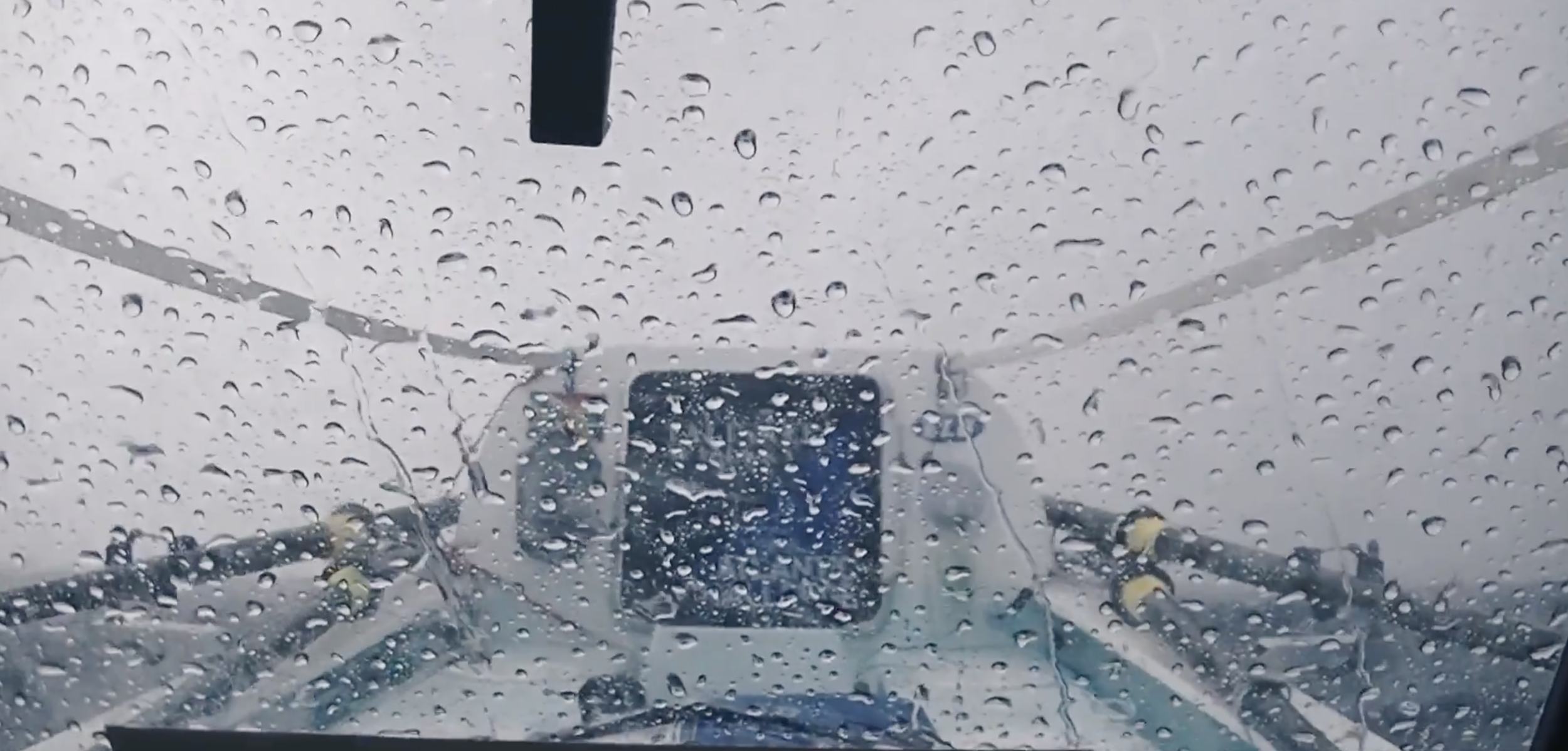 View through a rain-covered airplane cockpit window showing the airplane's front landing gear and the rainy sky.