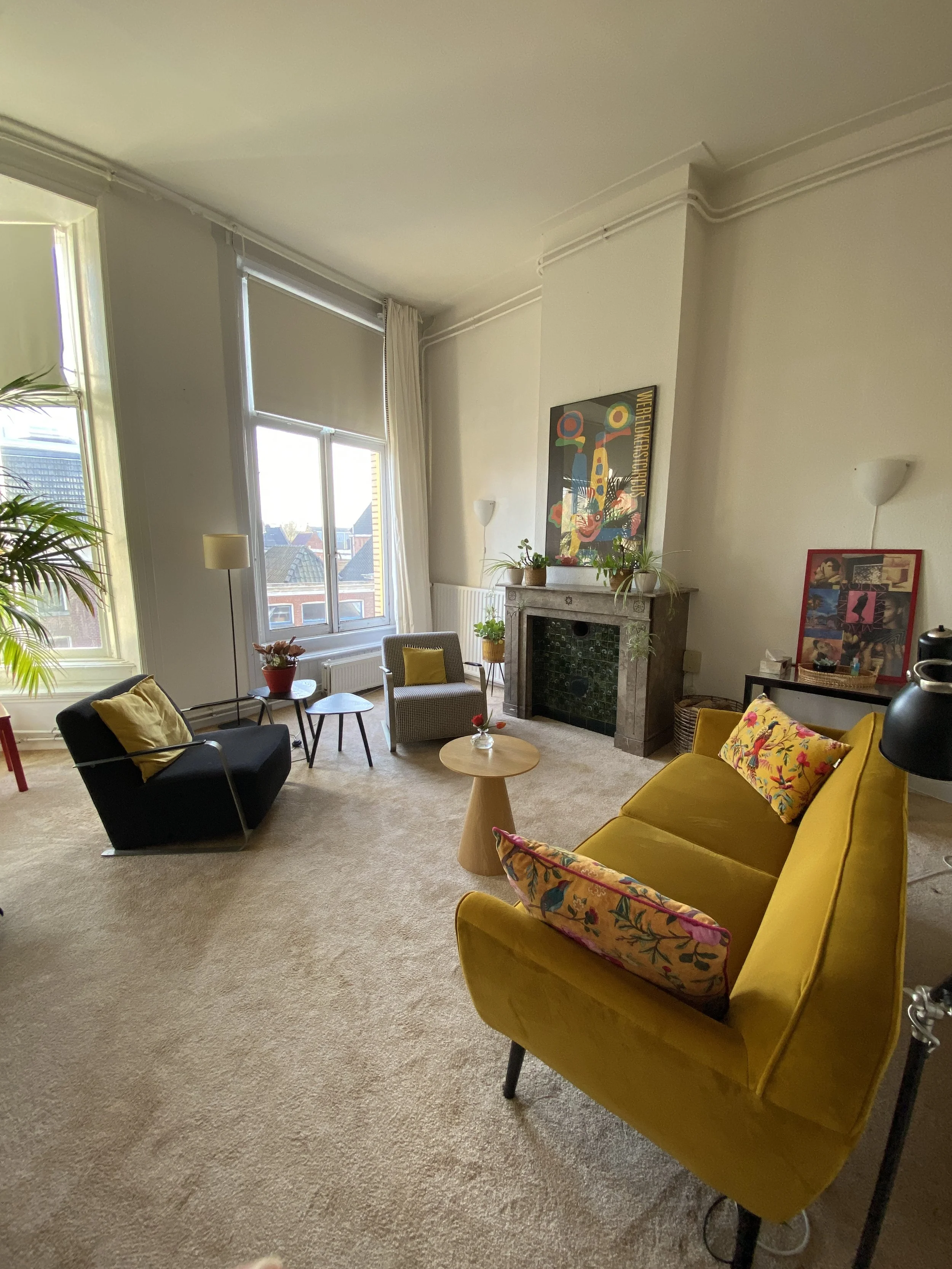 Living room with a yellow sofa, black chair, coffee table, potted plants, fireplace, artwork, and large windows with blinds and curtains.
