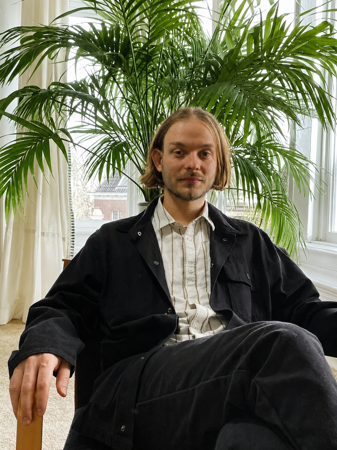 A young man with shoulder-length blond hair and light skin sitting in a room with natural light, a large green plant behind him, and cream-colored curtains near a window.