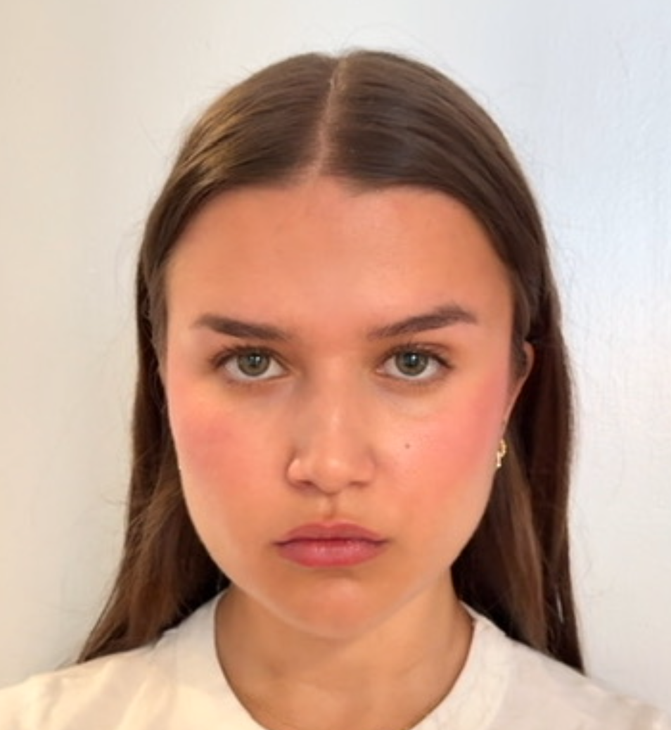 Close-up of a young woman with brown hair, blue eyes, wearing earrings and a light-colored shirt, standing against a plain light wall.