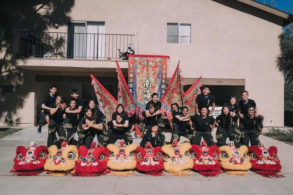Group of lion dancers dressed in black in front of colorful lion dance costumes. Team banners in fill up the background celebrating their team and cultureb