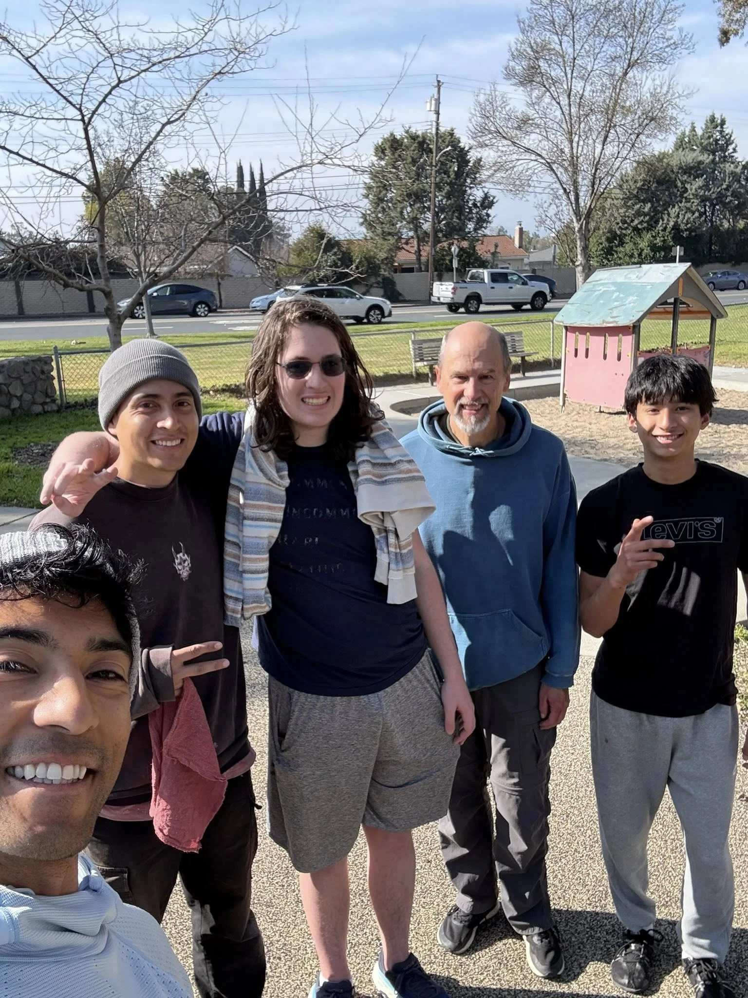 A group of five people — part of the parkour Sqvadron community — standing together outdoors in a park, smiling at the camera. There are trees, a playground, and a street with cars in the background.