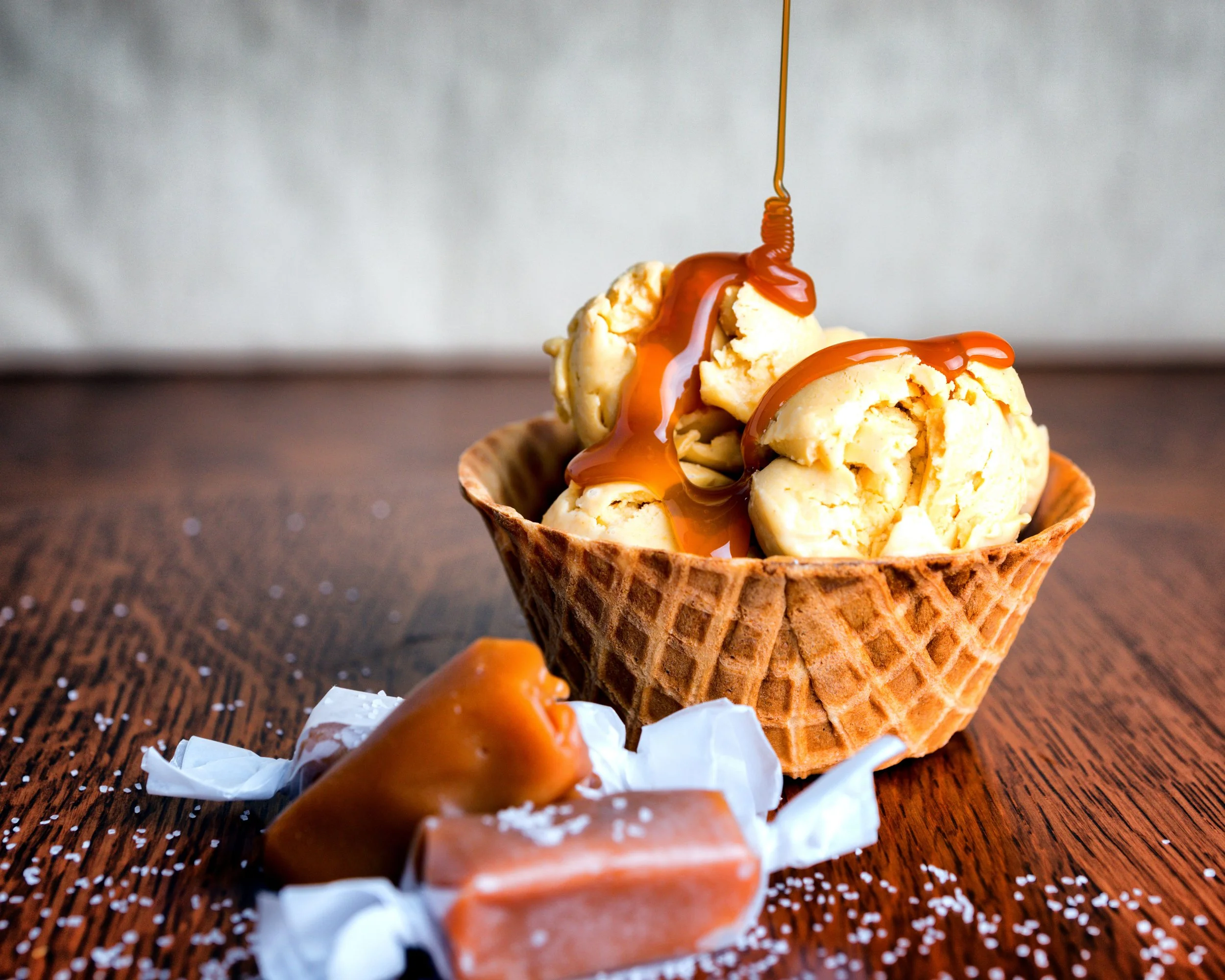 Scoops of vanilla ice cream topped with caramel sauce in a waffle cone, on a wooden surface with whipped cream and caramel candy in the foreground.