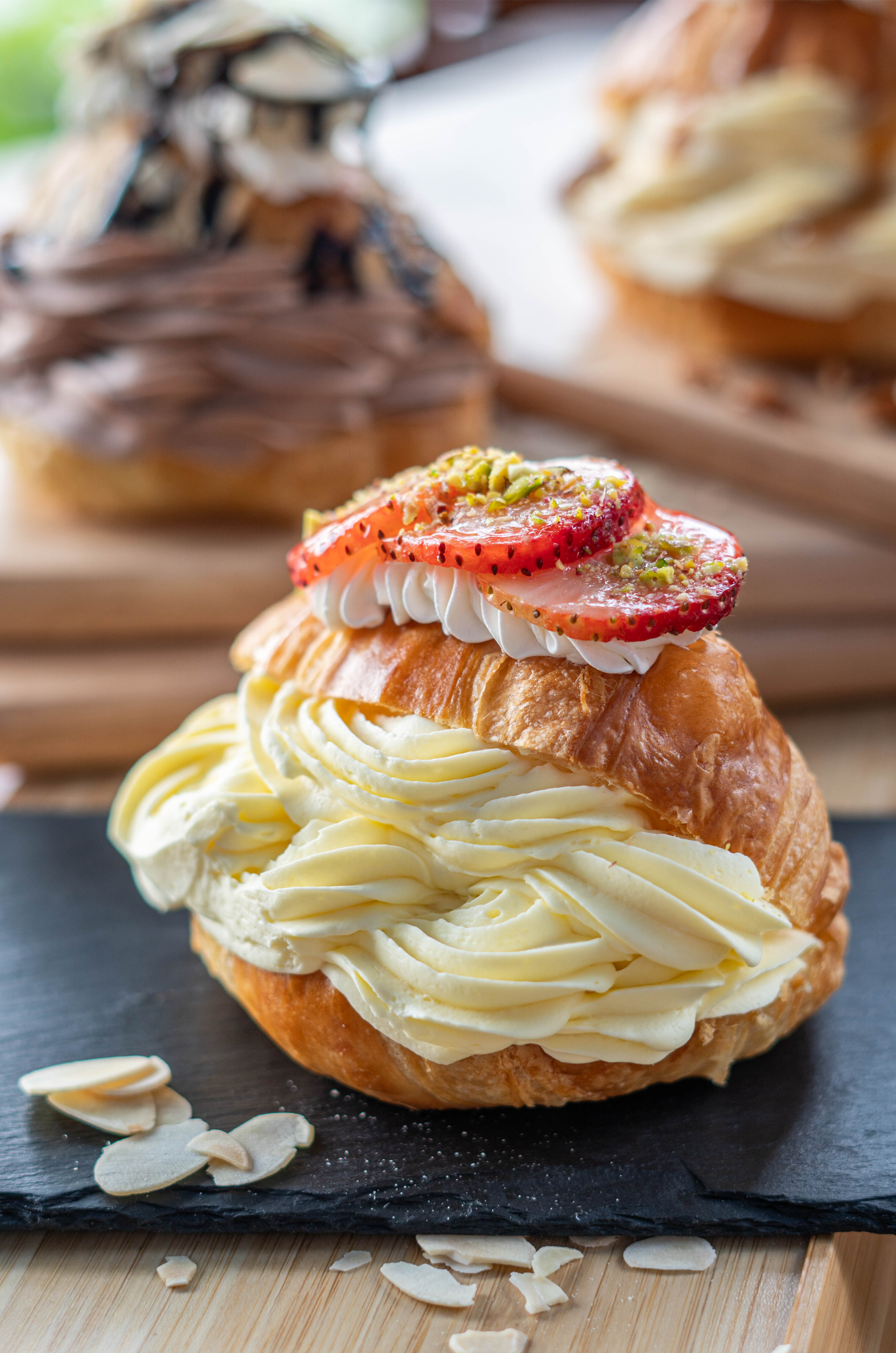 Close-up of a cream puff pastry topped with sliced strawberries and chopped pistachios on a dark slate serving board.