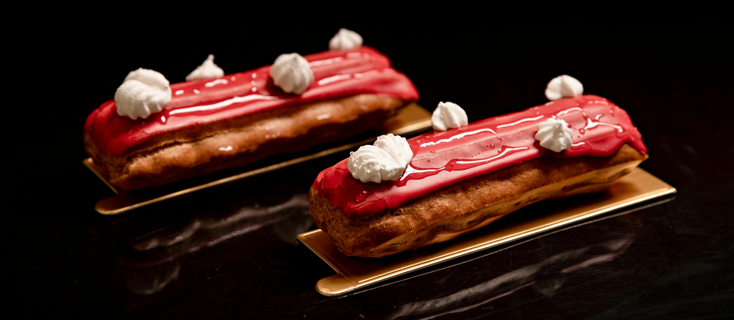 Two éclairs with pink frosting and white whipped cream dollops on top, placed on gold trays against a black background.