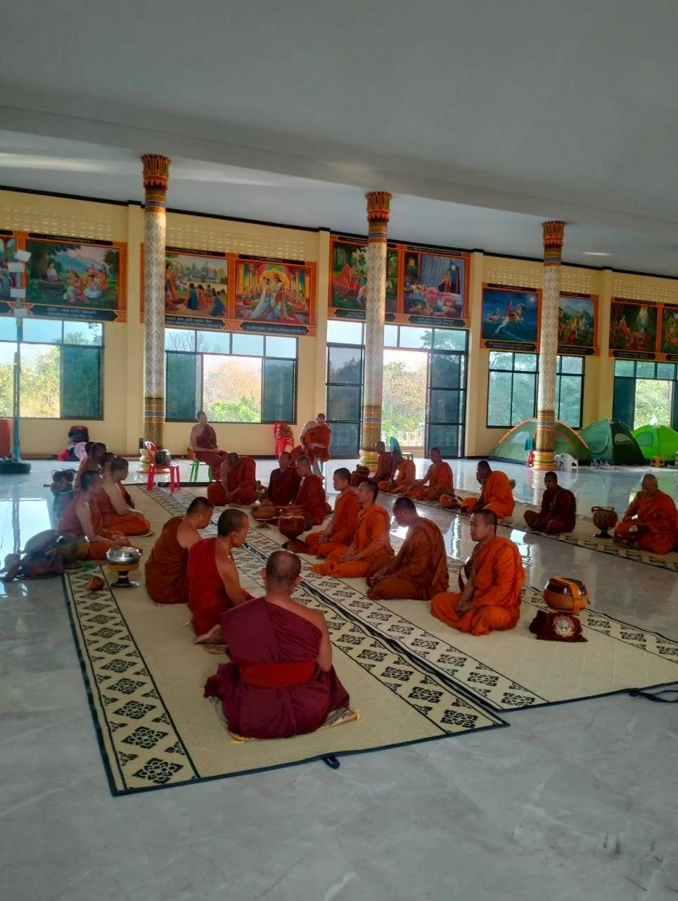 Group of monks sitting on a prayer mat inside a Buddhist temple, with colorful murals and large windows in the background.