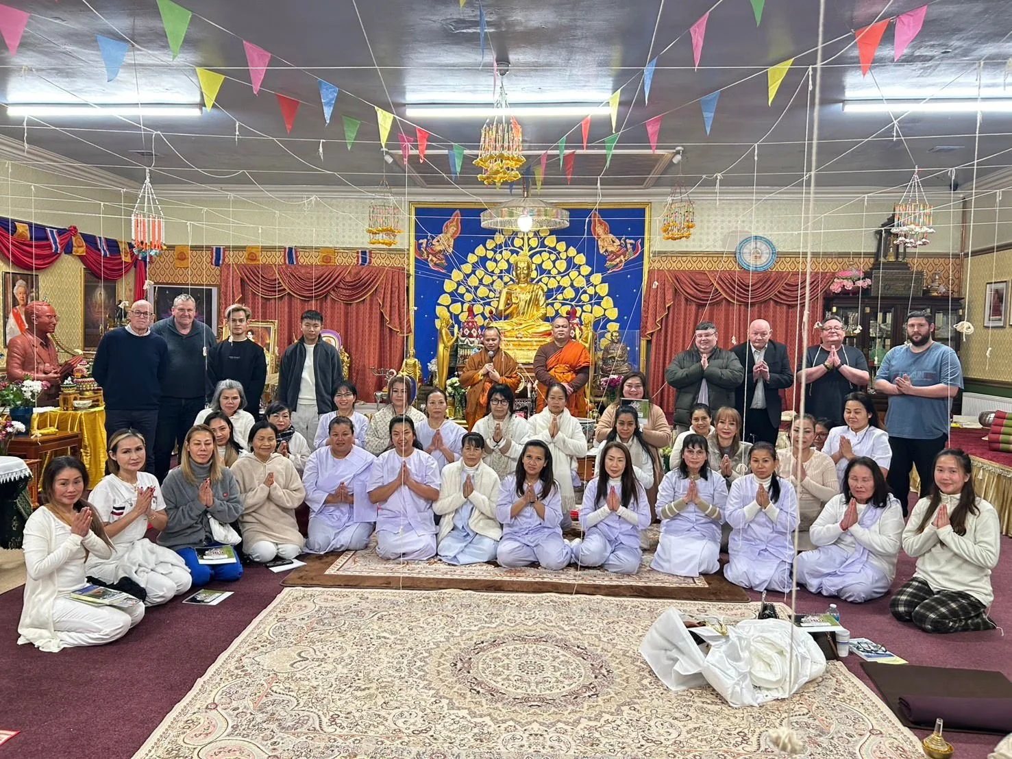A large group of people gathered in a Buddhist temple, sitting and standing in front of a gold Buddha statue on a blue backdrop. Some individuals are praying with hands in prayer position, others are standing or sitting with smiling faces. The temple