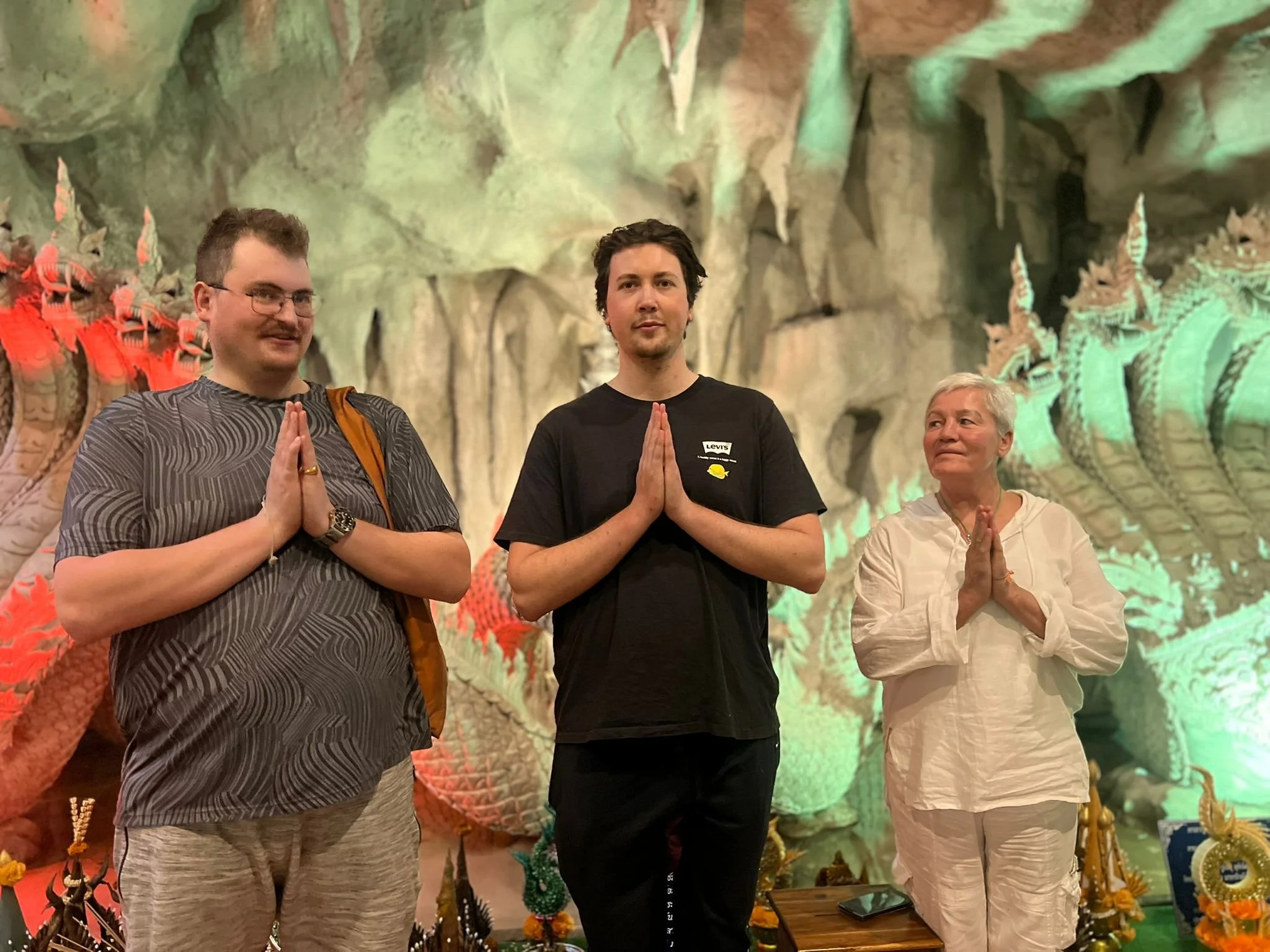 Three people standing with their hands together in a prayer position, inside a cave decorated with intricate dragon sculptures and colorful lighting.