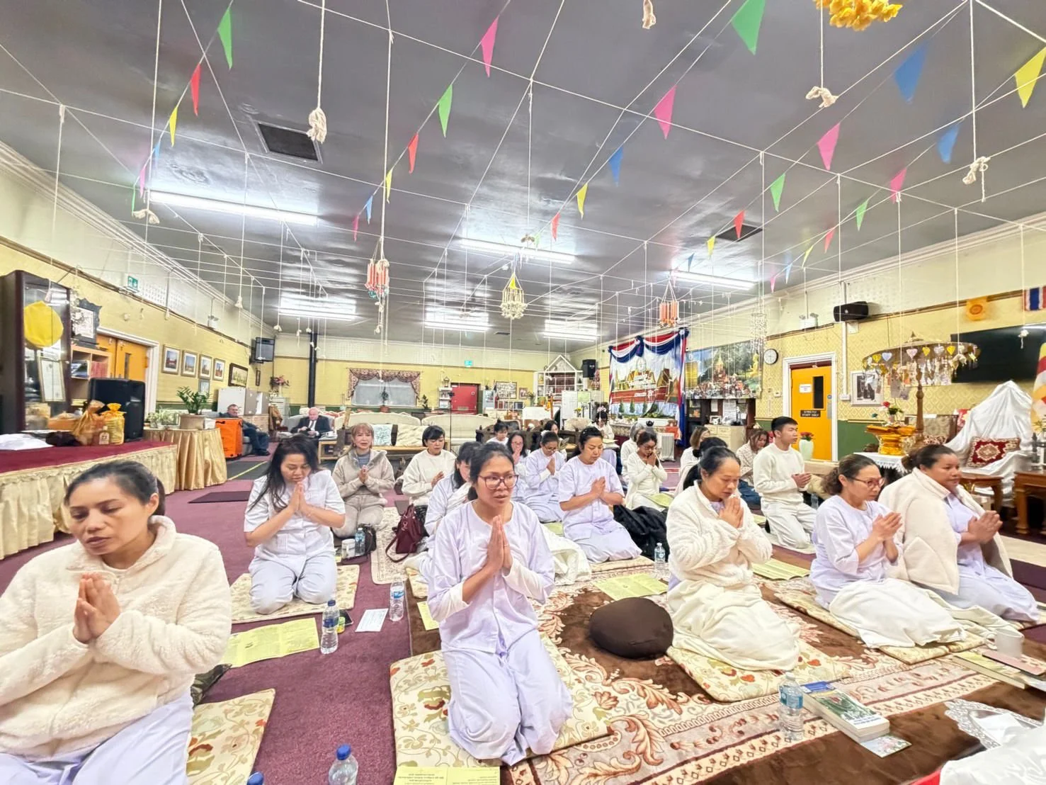 People participating in a spiritual or religious meditation or prayer session indoors, seated on cushions with hands pressed together in prayer pose, surrounded by decorative banners and religious artifacts.