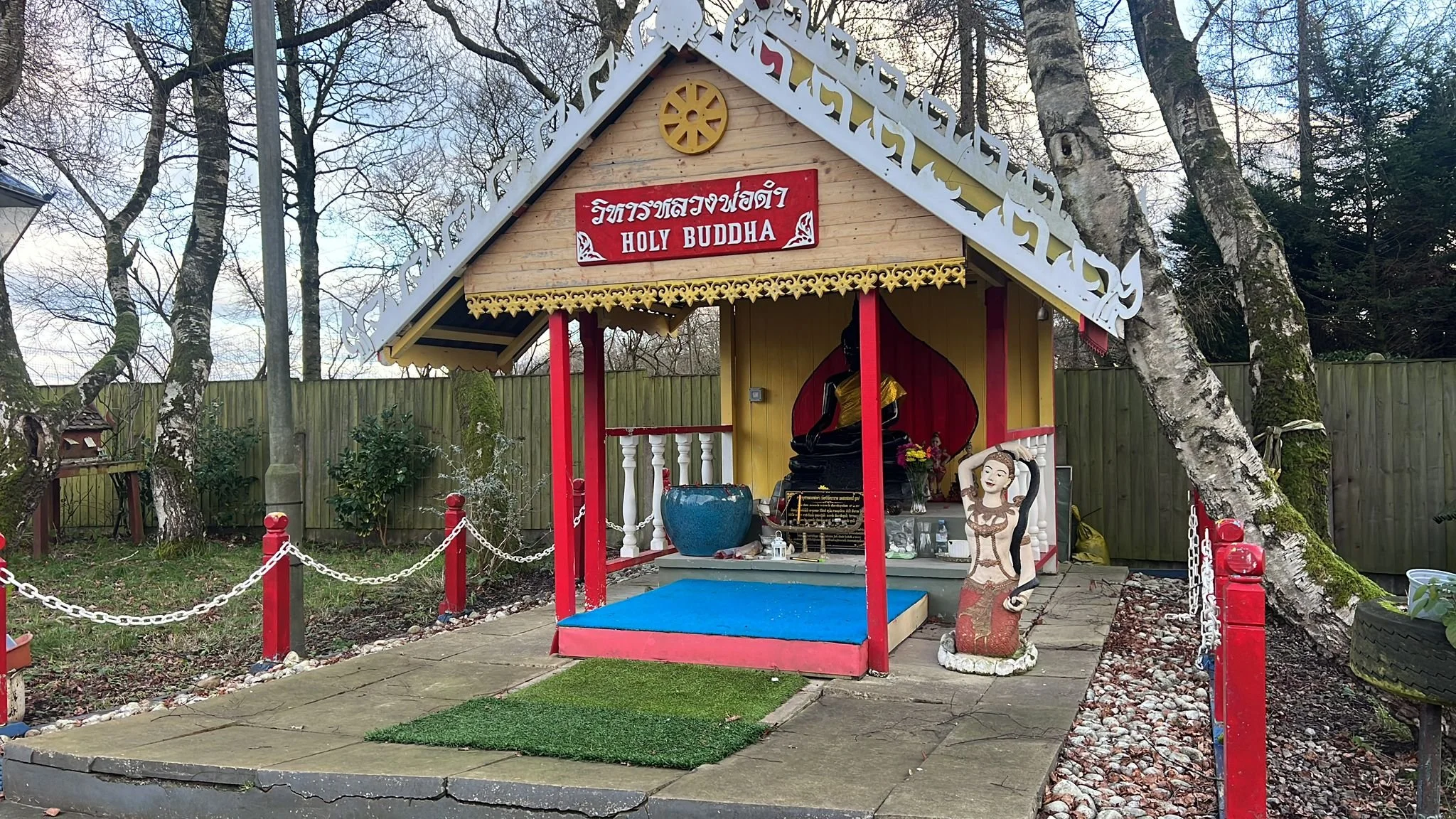 Small outdoor shrine dedicated to Buddha featuring a seated black Buddha statue, a golden statue of a woman, and a figure of a woman with arms raised. The shrine has a traditional Thai-style roof with ornate white and gold trim and a red sign with Th