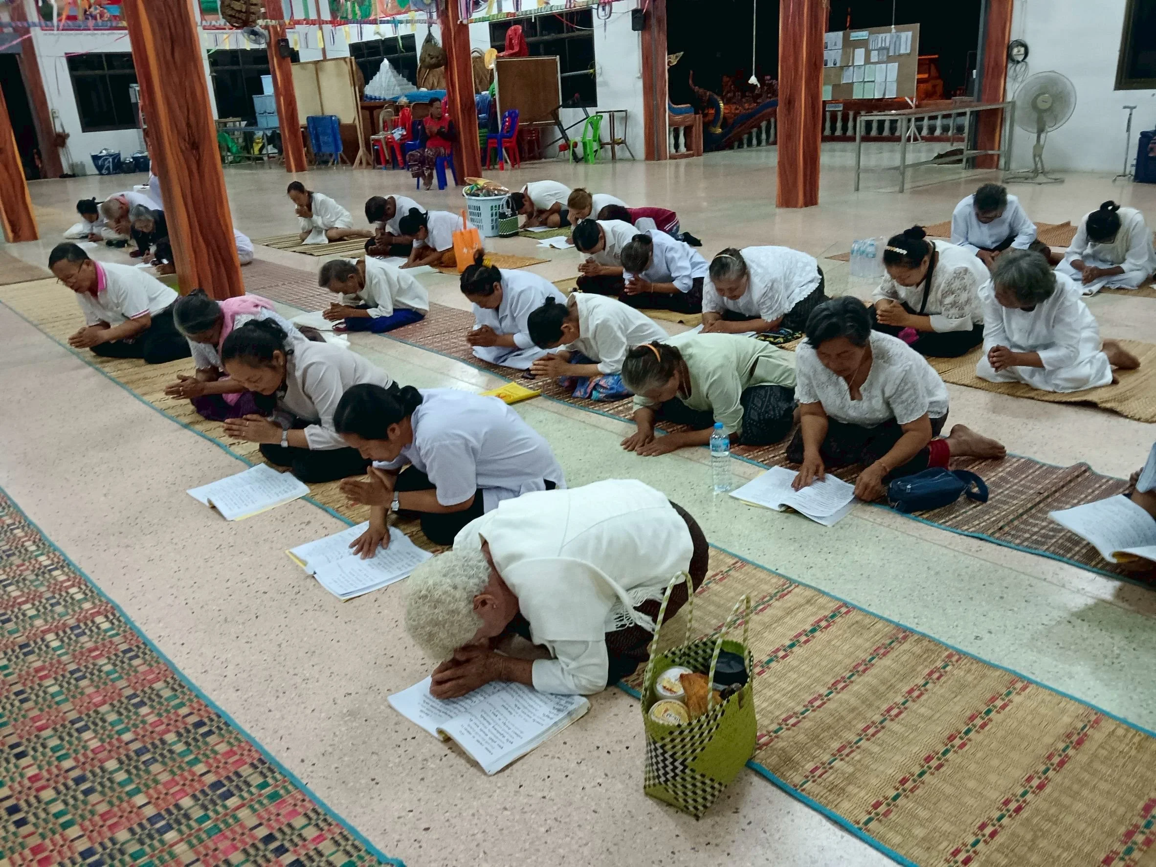 People gathered on the floor in a large hall, participating in a prayer or meditation session, sitting on mats with papers and drinks in front of them.